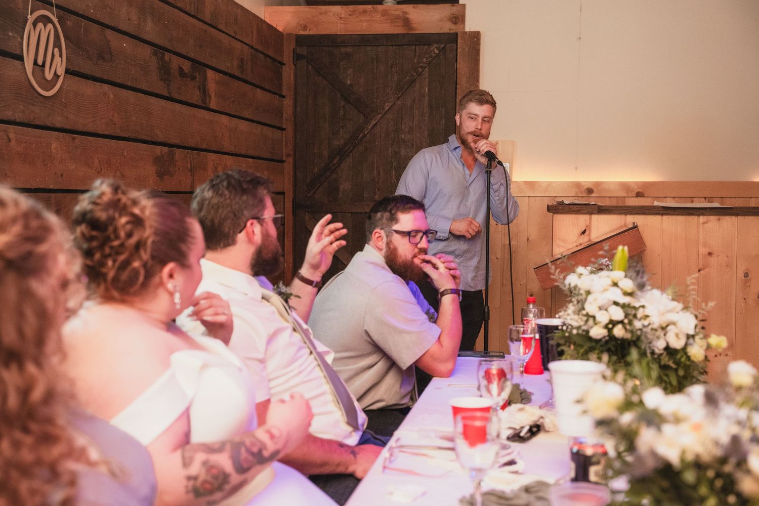 A man gives a heartfelt wedding speech at a microphone while guests listen at decorated reception tables.