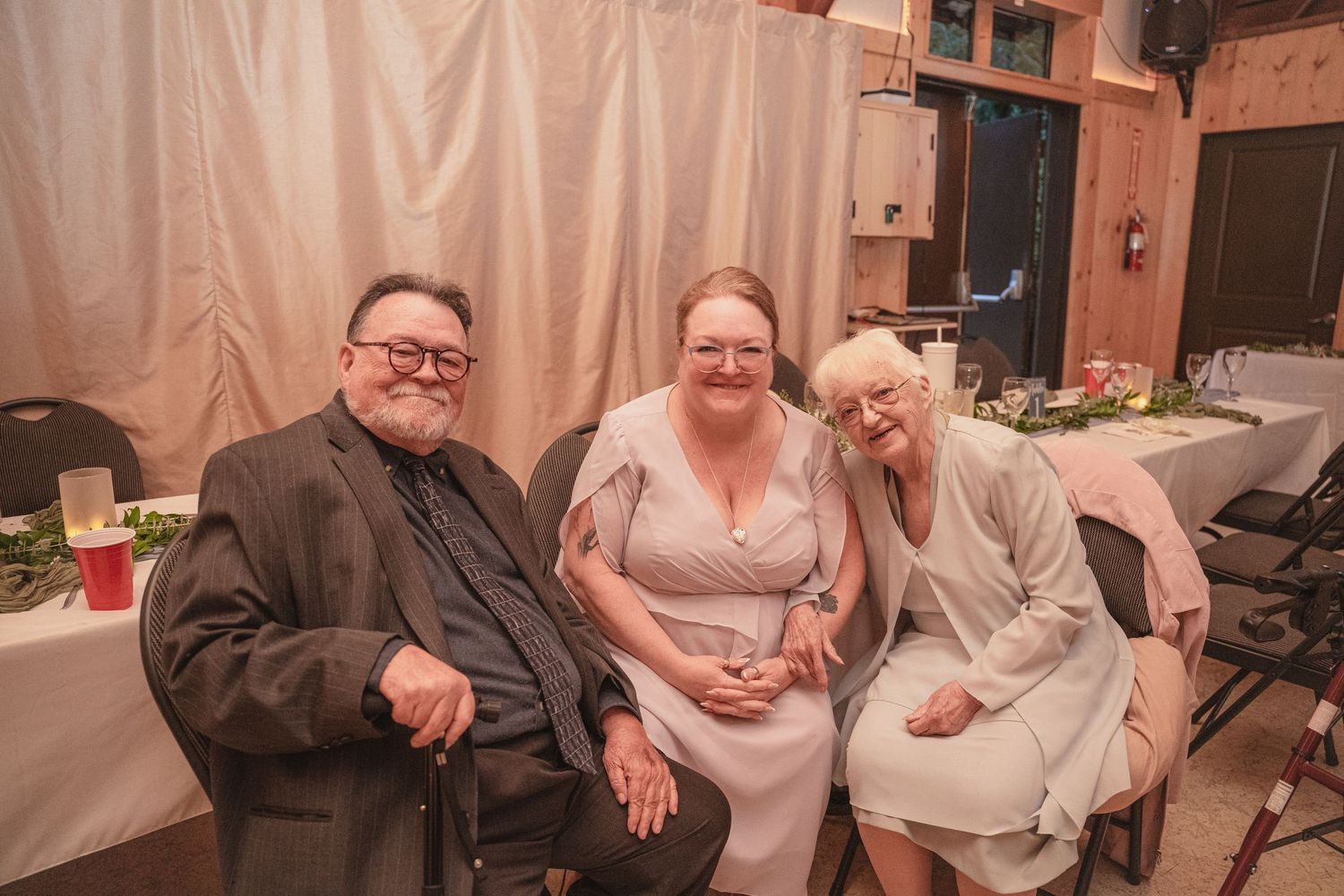 Three smiling wedding guests pose together at a rustic reception venue during a wedding celebration.