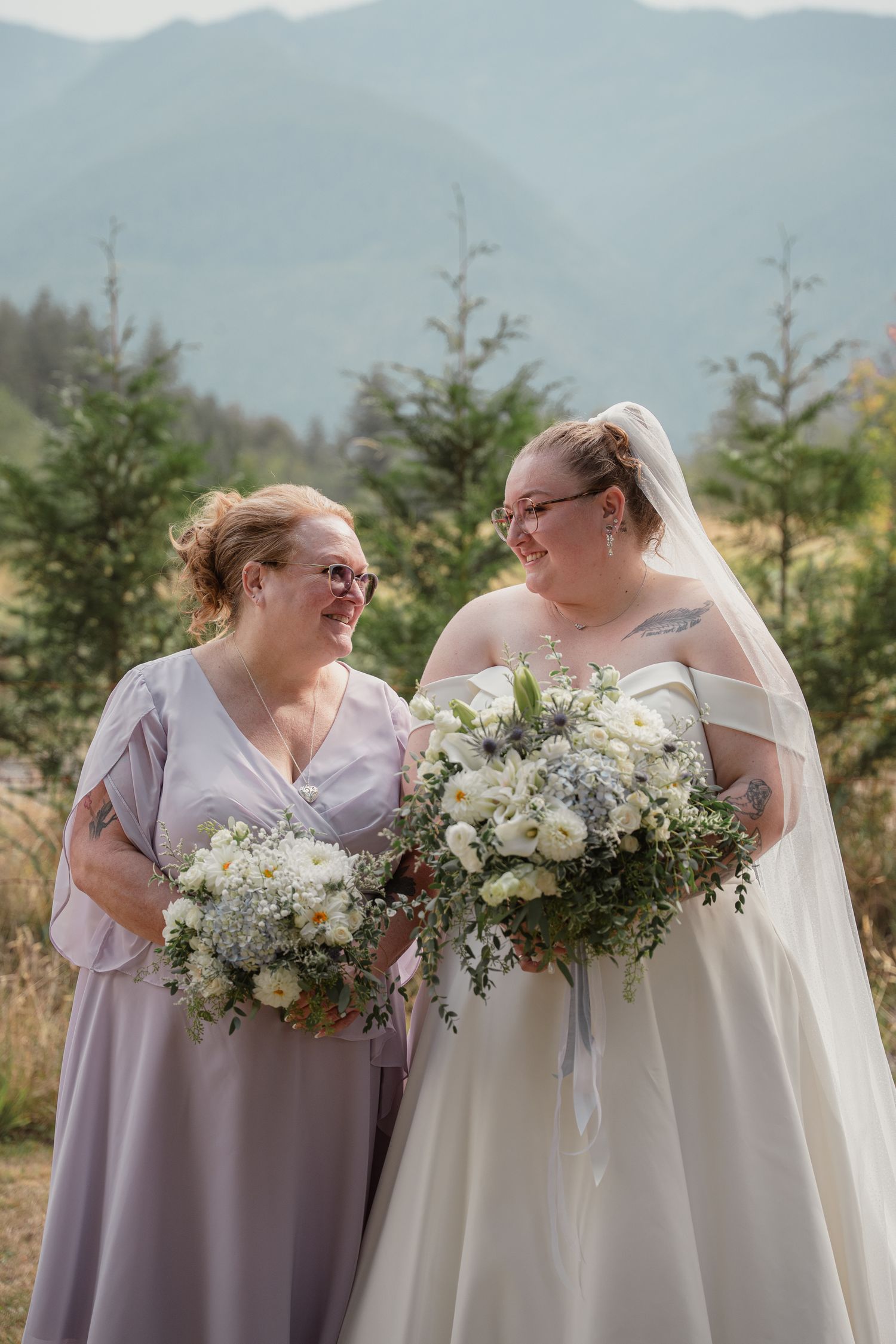 Bride in white gown and veil holds bouquet beside bridesmaid in lavender dress, both smiling, with mountain backdrop.