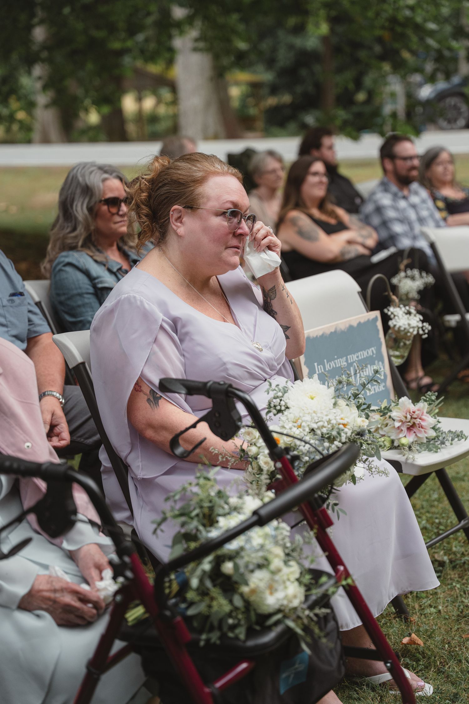 Emotional wedding guest wipes tears while seated beside a memorial chair with flowers at outdoor ceremony.