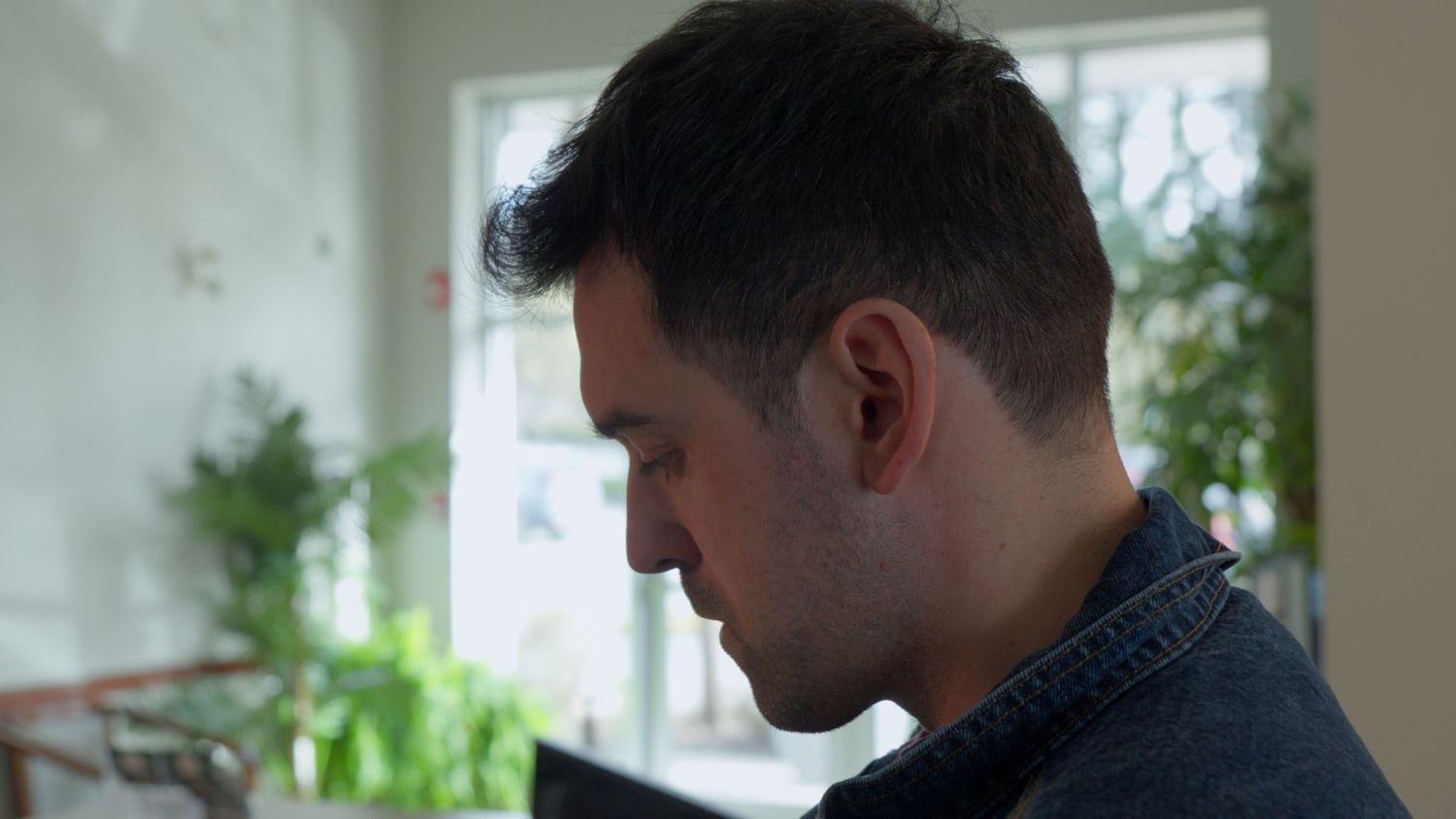 Close-up profile of a young man with short dark hair wearing a denim jacket, looking down indoors.
