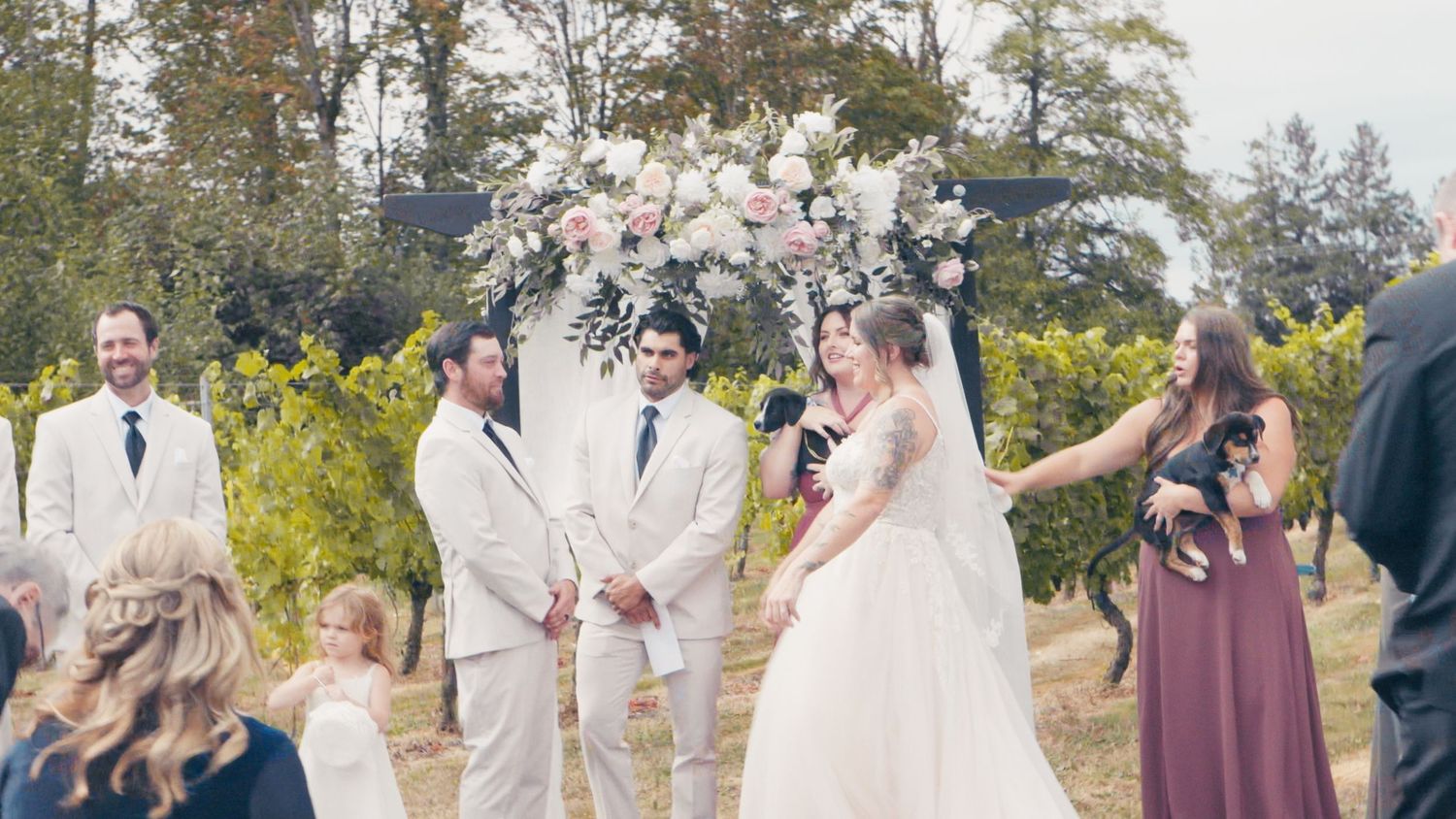 Joyful outdoor wedding ceremony at a vineyard with couple exchanging vows under a floral arch.