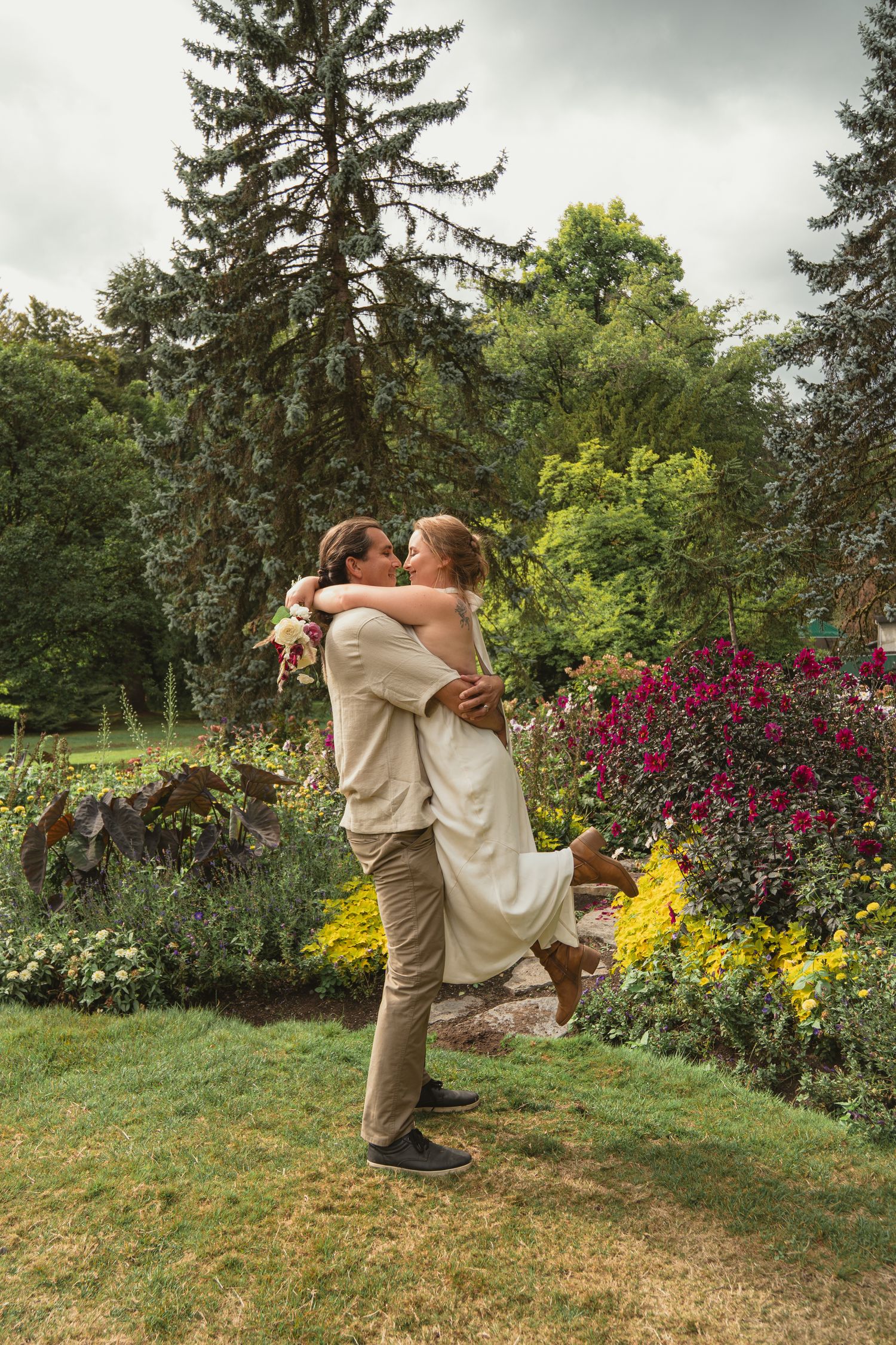 Groom lifting smiling bride in a white dress in a vibrant garden with tall evergreen trees.