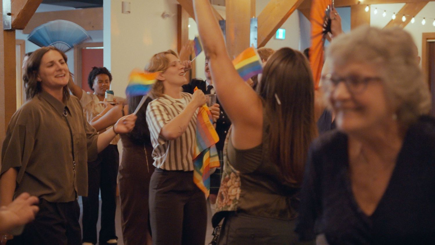 Wedding guests celebrate and wave Pride rainbow flags joyfully inside a rustic venue.