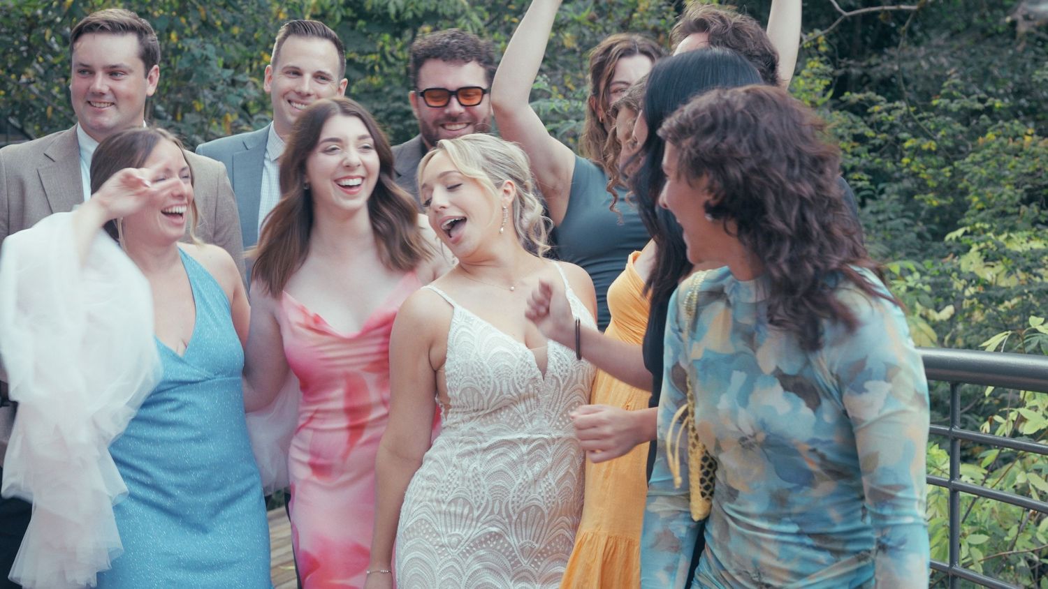 Bride in white wedding dress laughing joyfully with wedding guests outdoors in a garden setting.