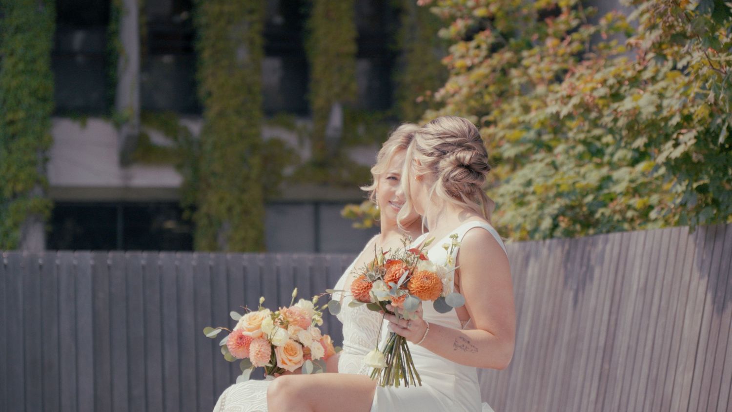 Two brides in white dresses sitting together holding beautiful orange and peach bouquets outdoors on their wedding day.