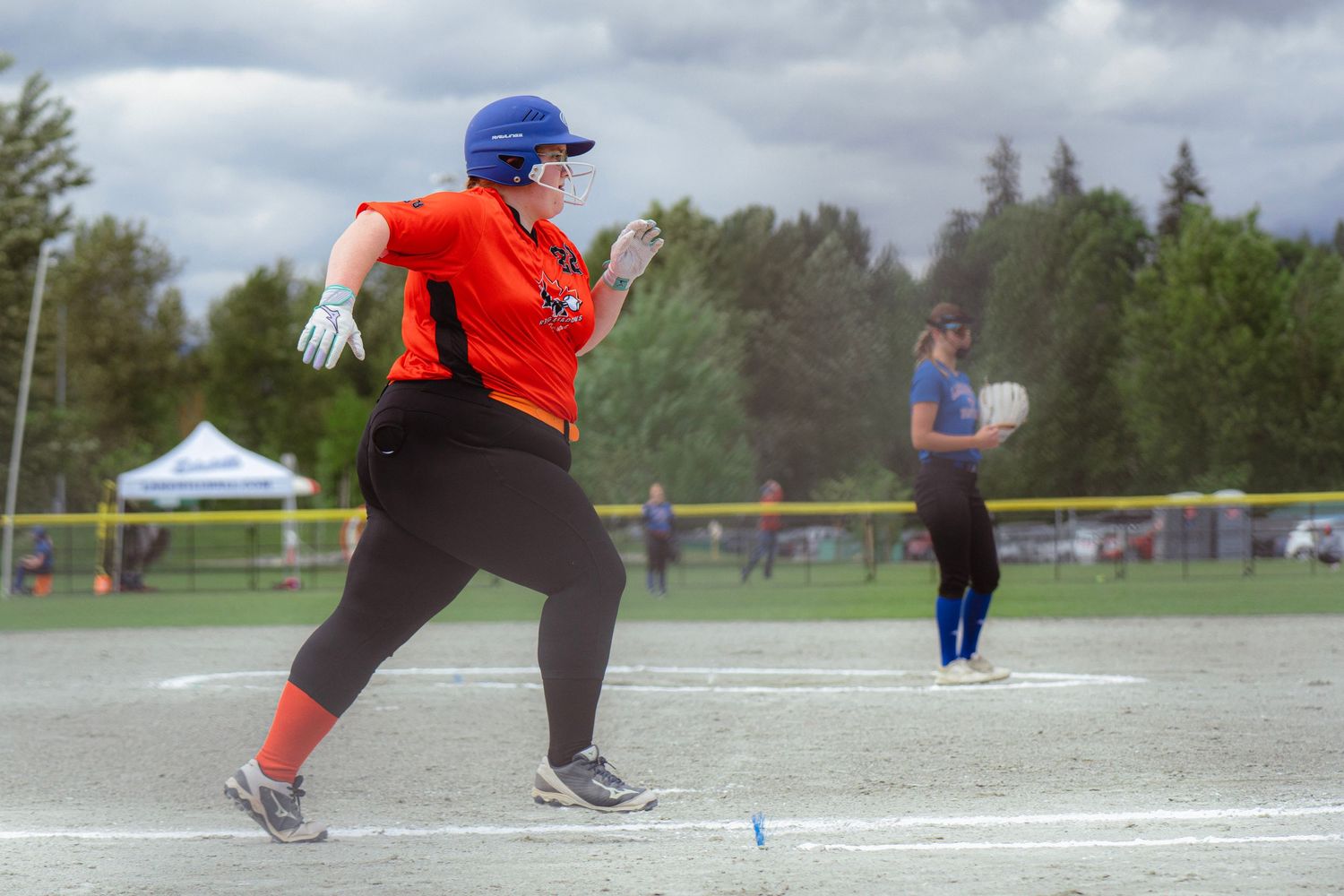 Female softball player in orange uniform running to first base during a softball game.