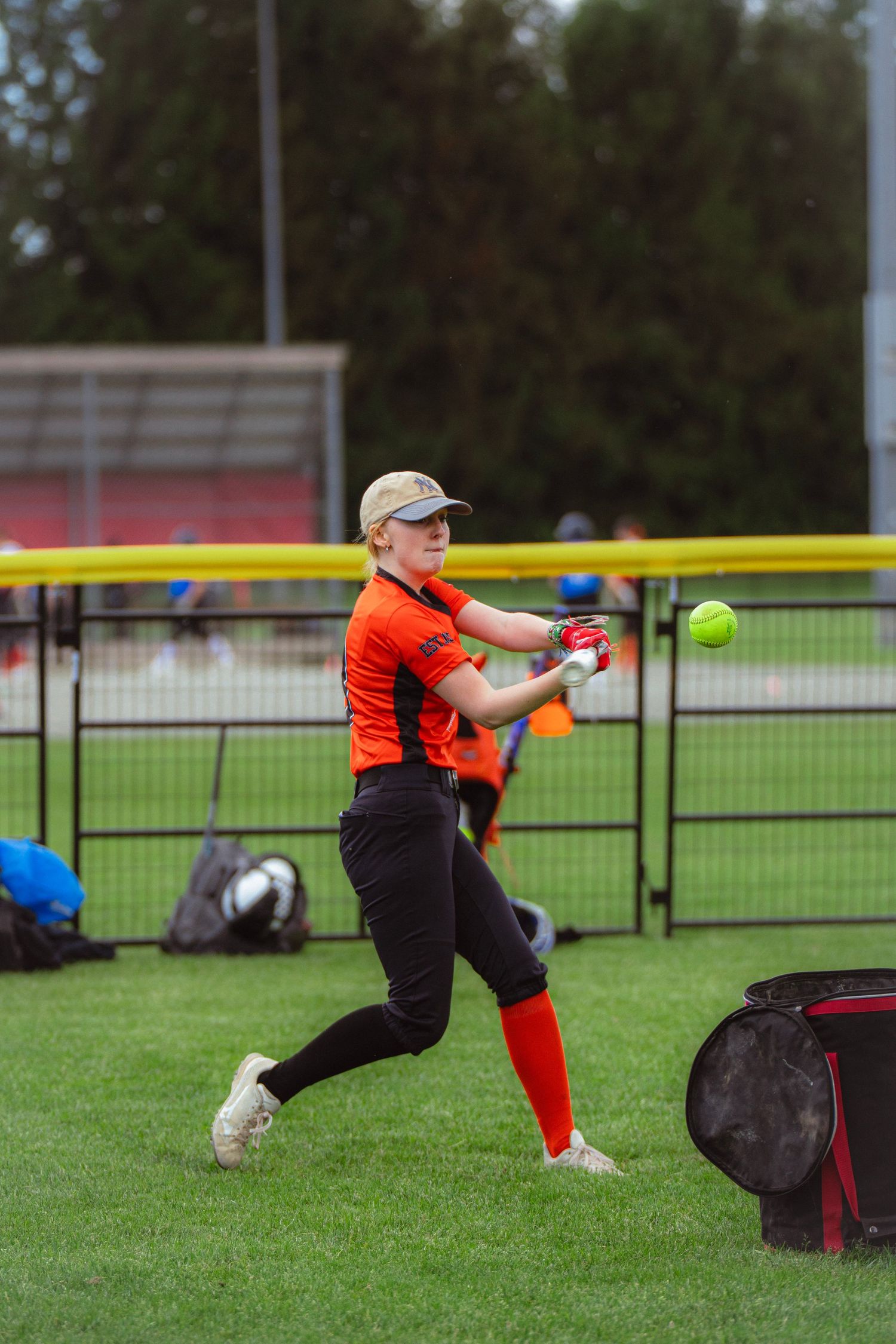 Female softball player in orange uniform making contact with yellow ball during batting practice.