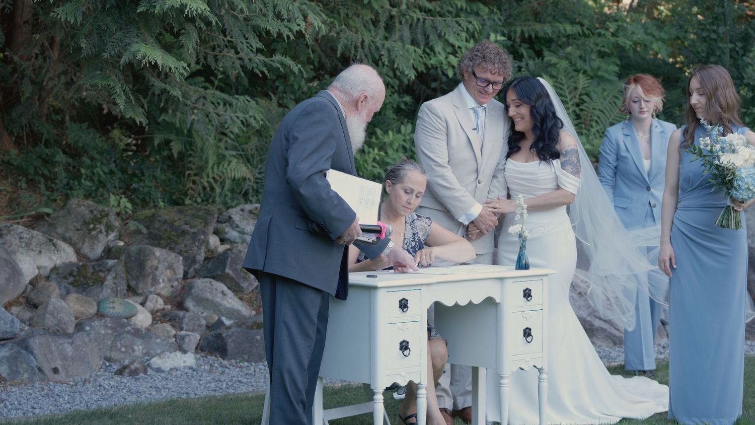 Wedding party gathers as bride signs marriage certificate at white desk during outdoor ceremony.
