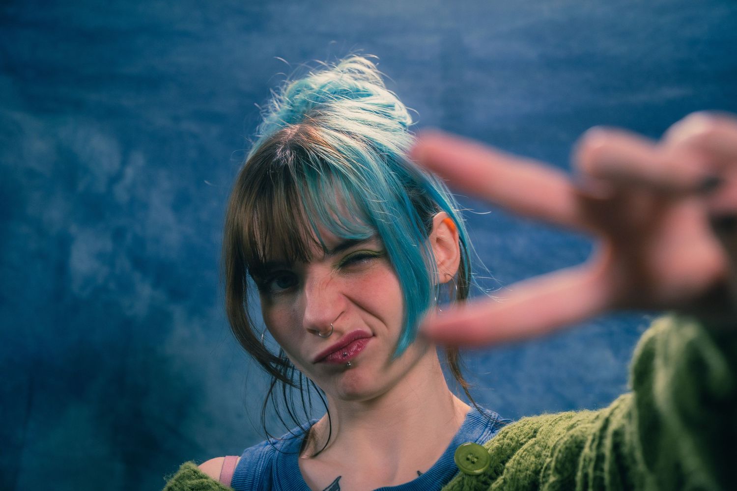 Young woman with blue hair making peace sign and playful expression in blue-toned studio photo.