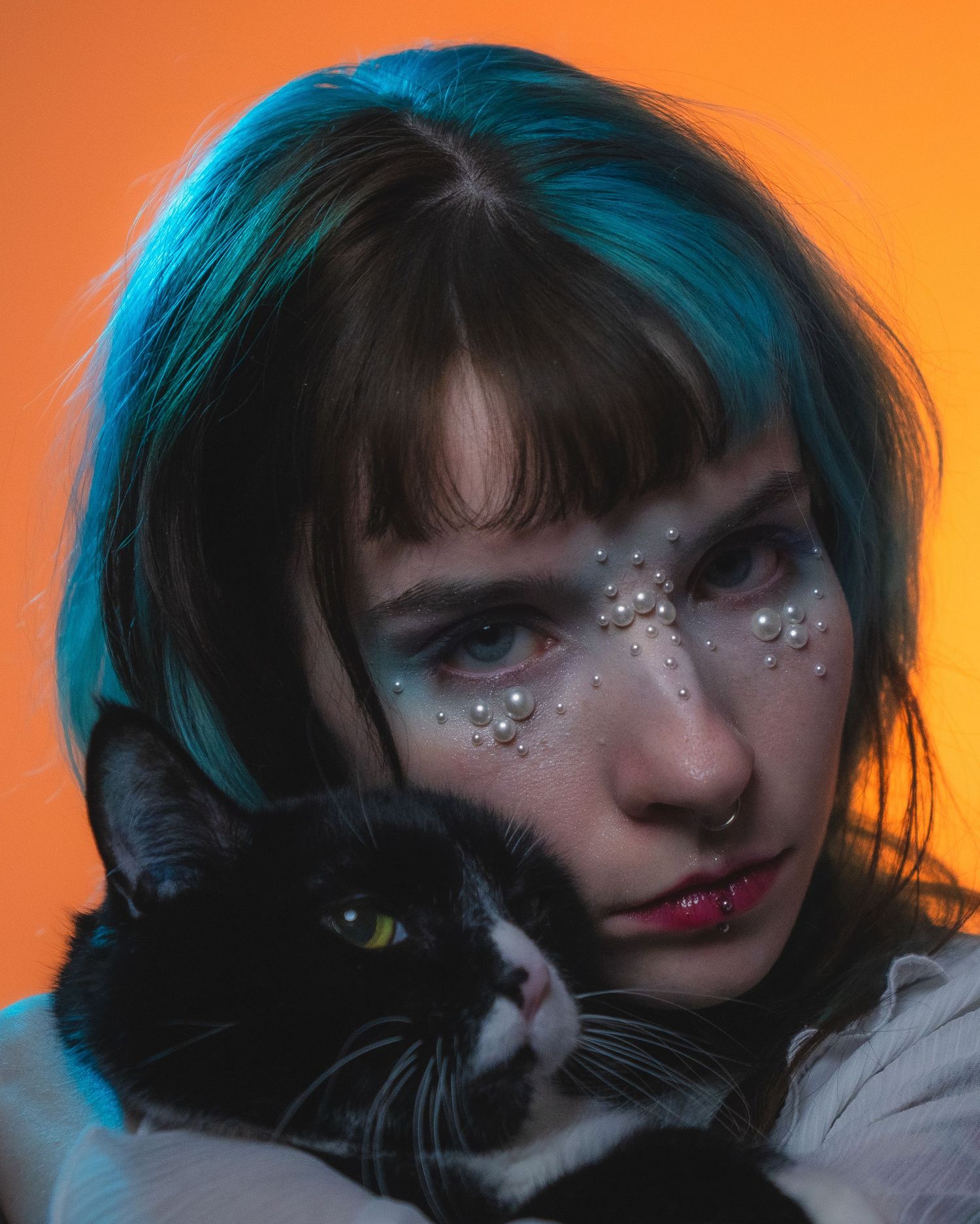 Close-up of woman with teal hair and pearl face gems cuddling a tuxedo cat on orange background.
