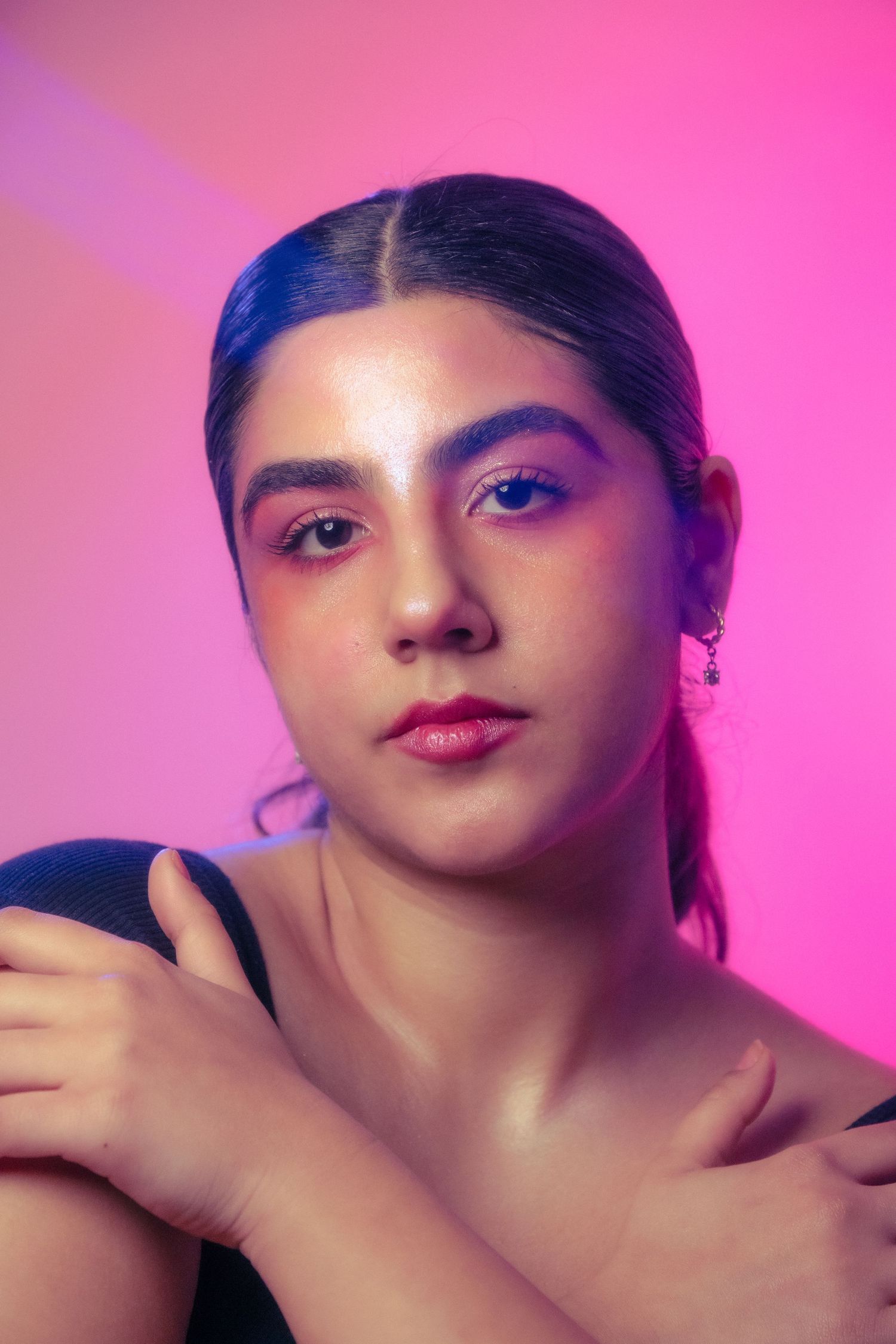 Young woman with pink eye makeup crossing arms over chest in a serious pose against a pink neon background.