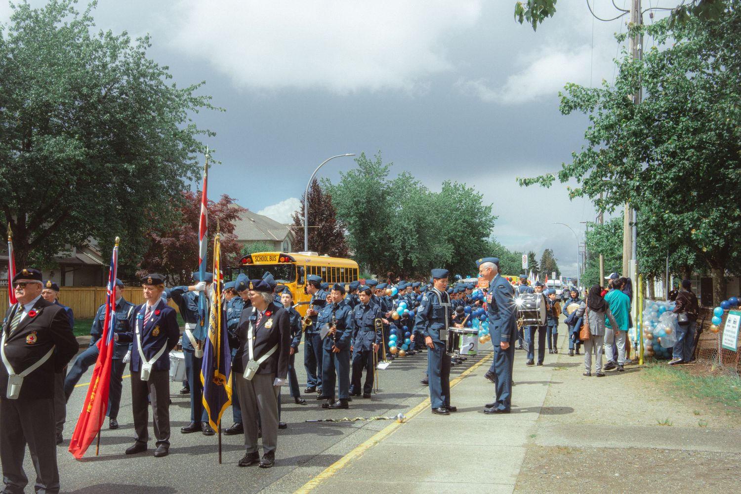 Military cadets and veterans assemble on a tree-lined street for a formal outdoor parade ceremony.