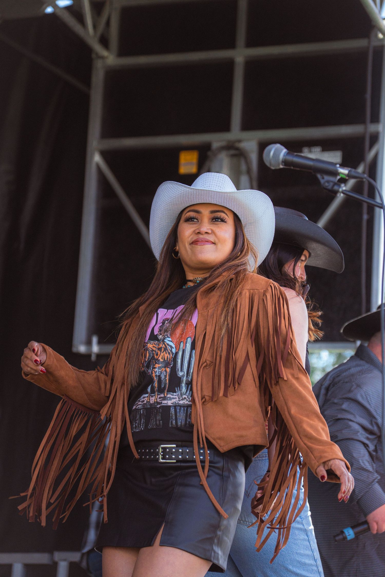 Energetic female performer in white cowboy hat and brown fringe jacket dances on outdoor festival stage.