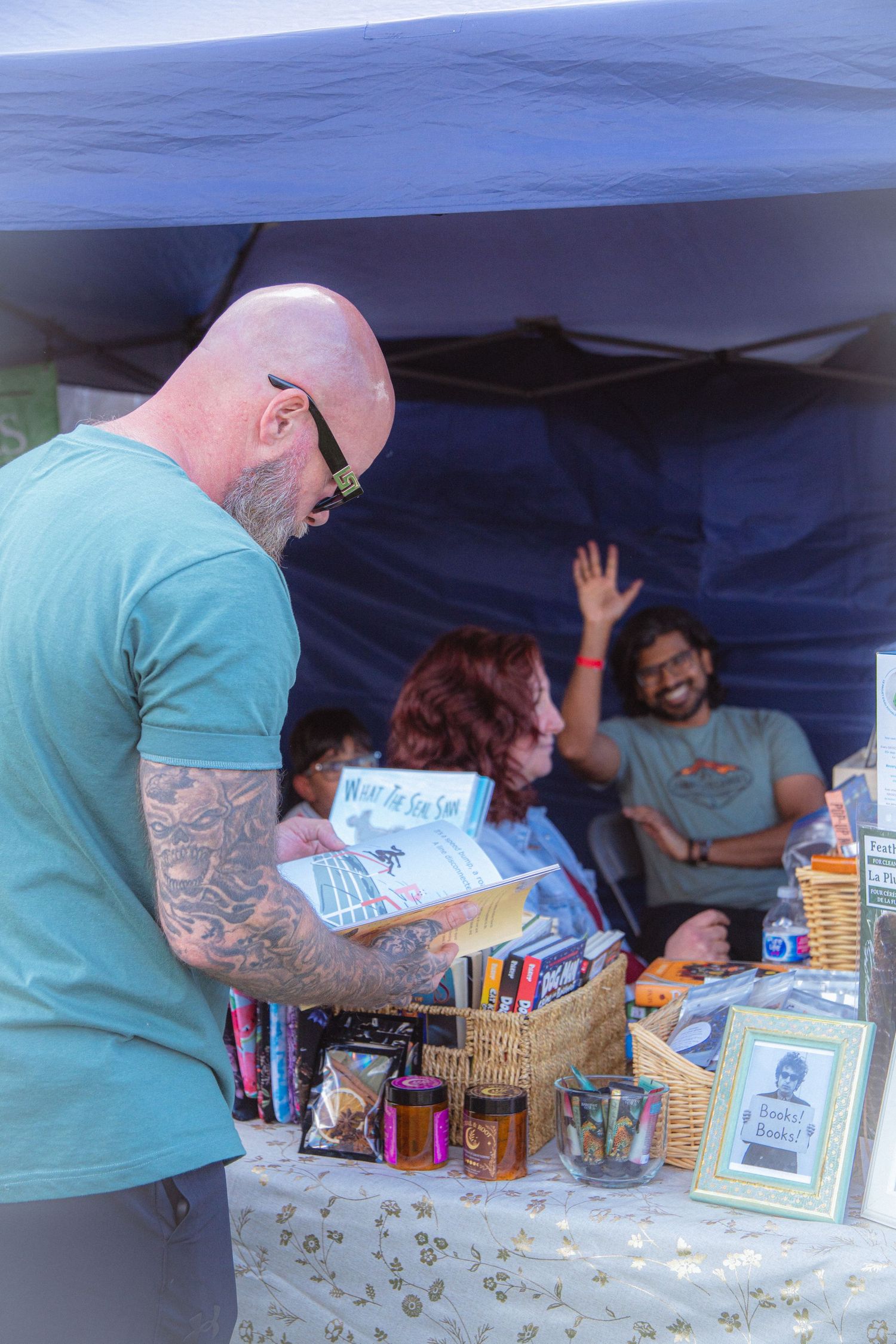 Tattooed bald man browses books and goods at an outdoor festival vendor booth.