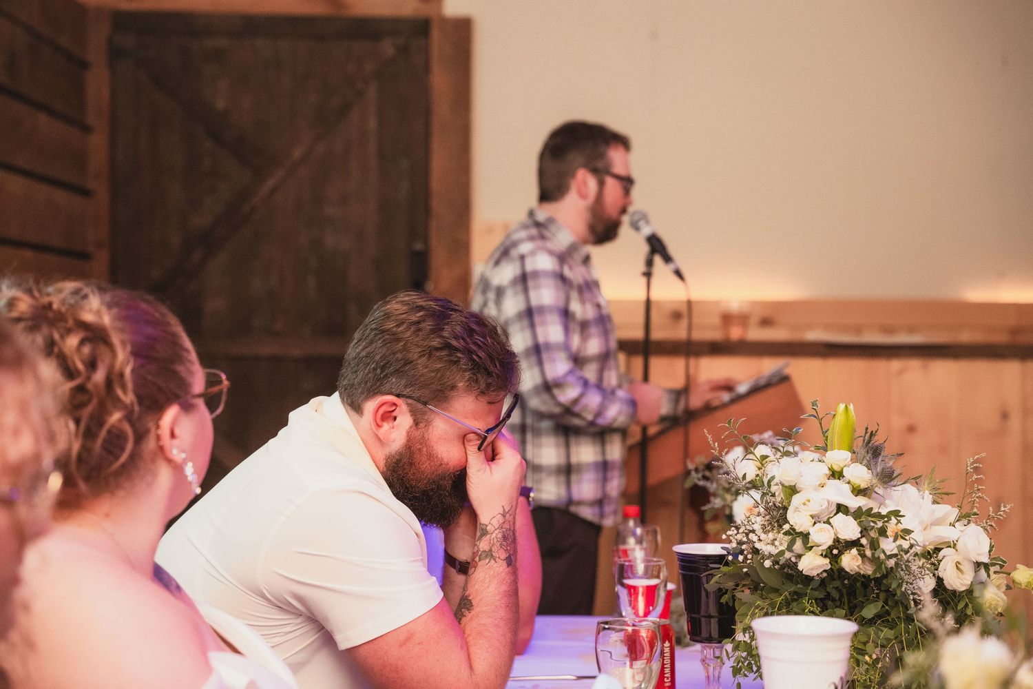 Emotional man covers his face during a heartfelt wedding speech in a rustic barn reception venue.
