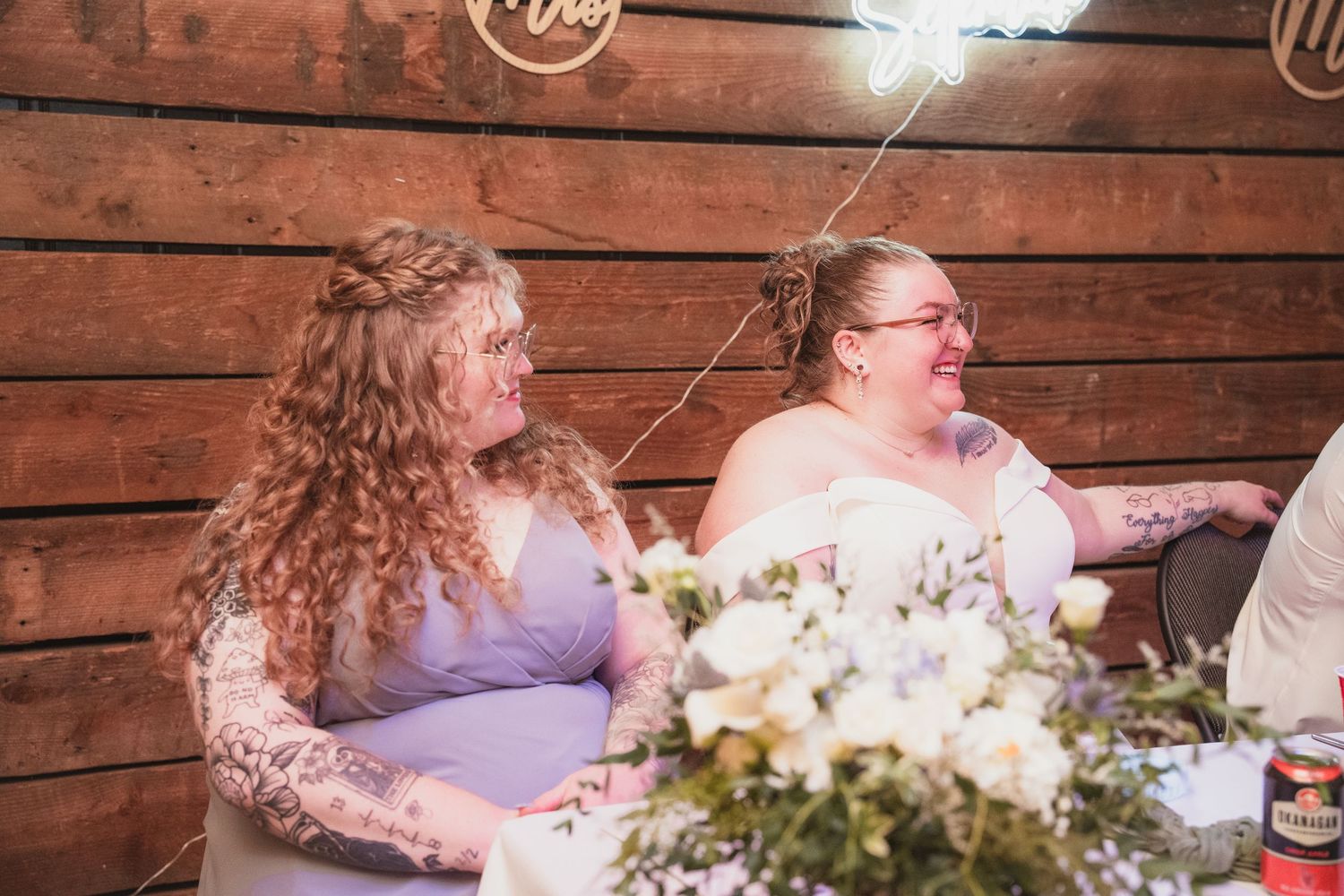 Bride and bridesmaid in lavender dress laugh emotionally during wedding reception speeches at barn venue.