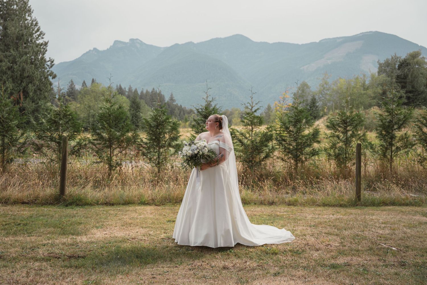 Bride in white gown and cape holding bouquet stands in scenic mountain meadow with evergreen trees backdrop.