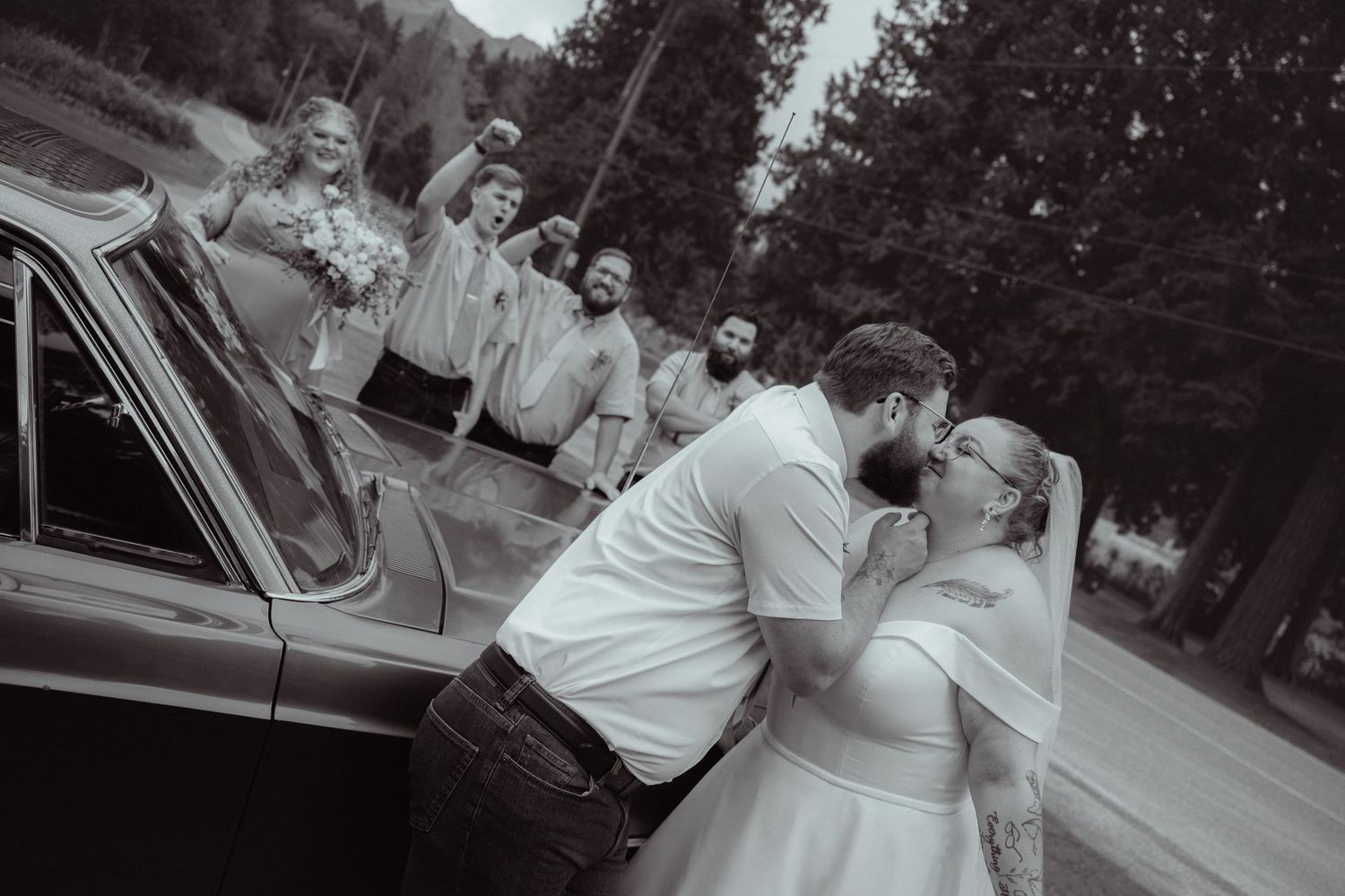 Bride and groom share kiss beside vintage car as wedding party celebrates behind them.