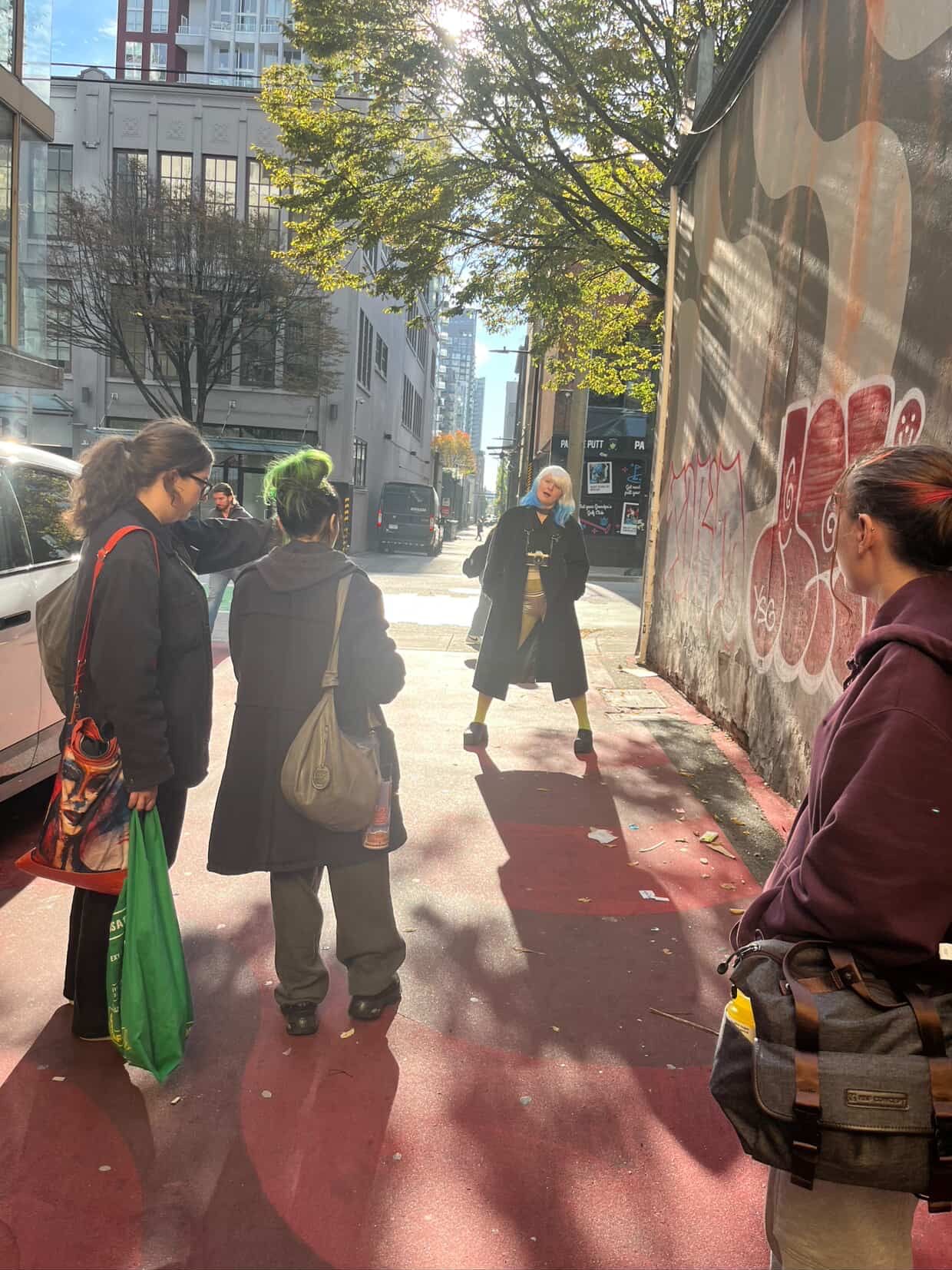 Small group gathered on a red urban sidewalk beside a graffiti-covered wall on sunny day.
