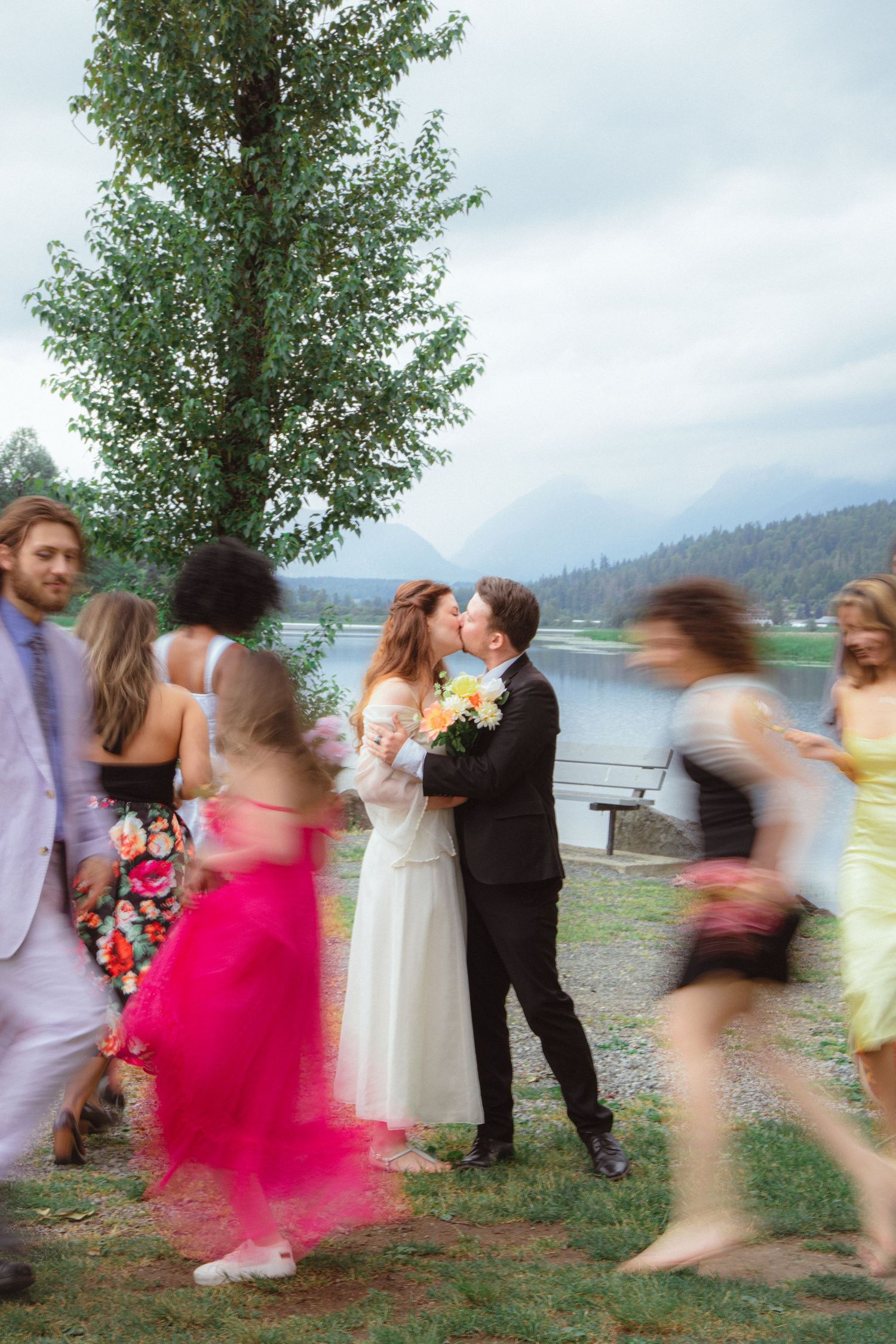 Bride and groom share a kiss while guests in colorful dresses move around them near a lake.