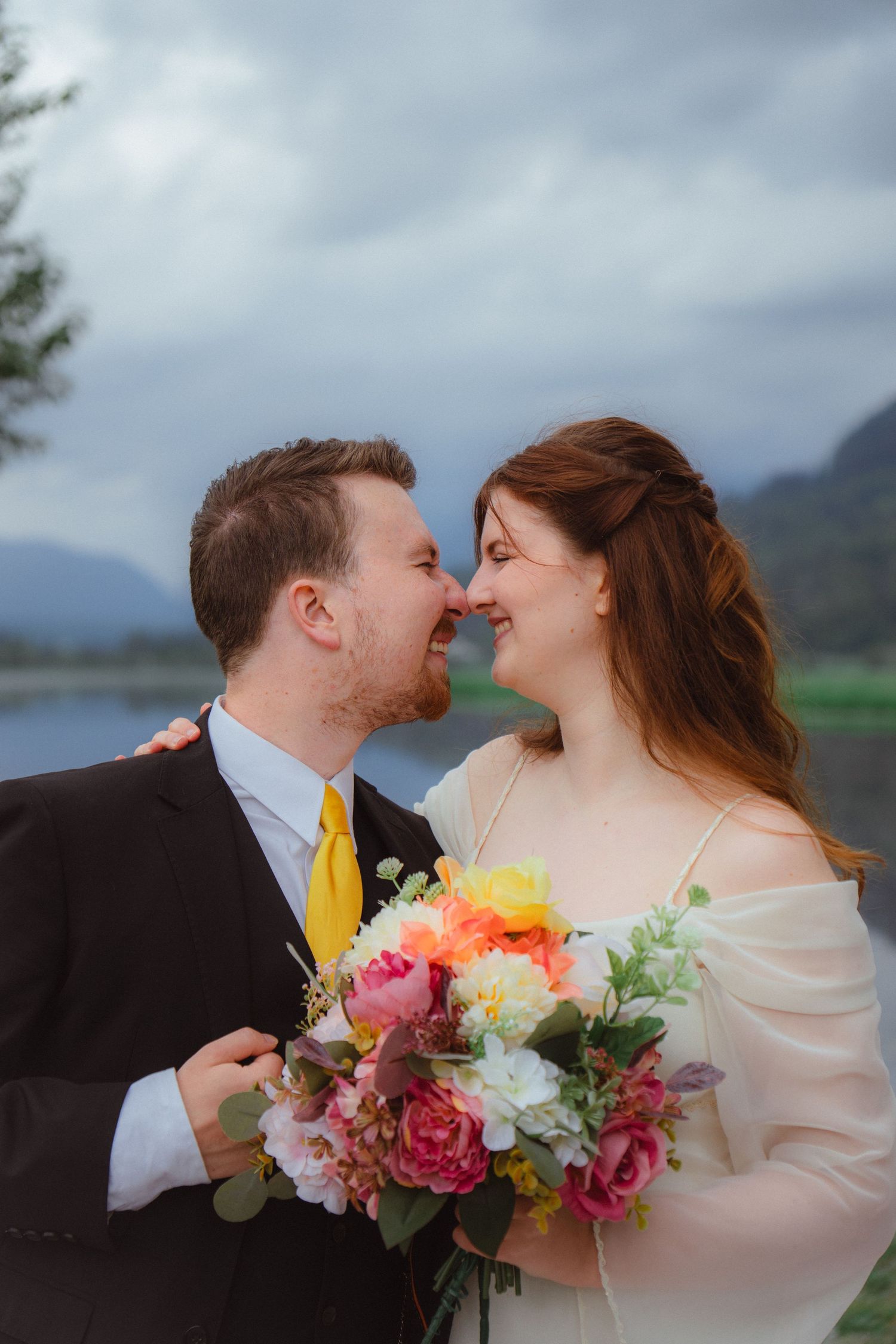 Laughing bride and groom nearly kiss while holding a colorful bouquet outdoors.