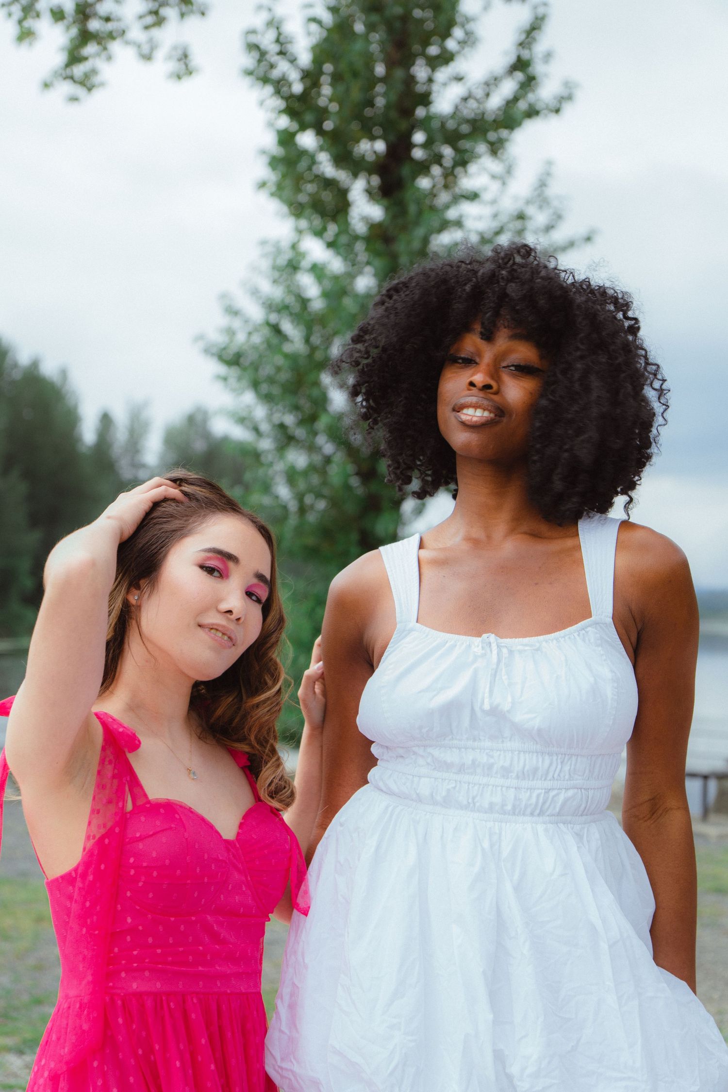 Two women in pink and white dresses smiling together outdoors near tall tree on cloudy day.