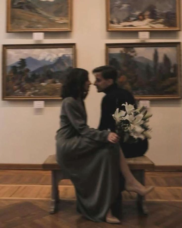 Couple sharing a romantic moment on a bench in an art gallery, holding white lilies with landscape paintings on the wall.
