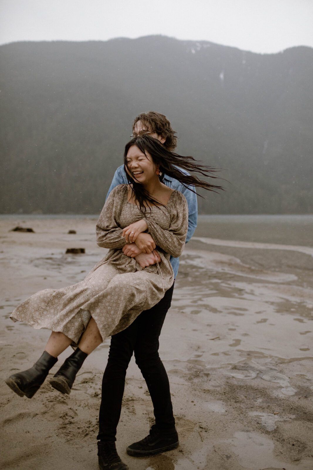 Couple laughing and spinning on a sandy beach with misty mountains in the background on a cloudy day.