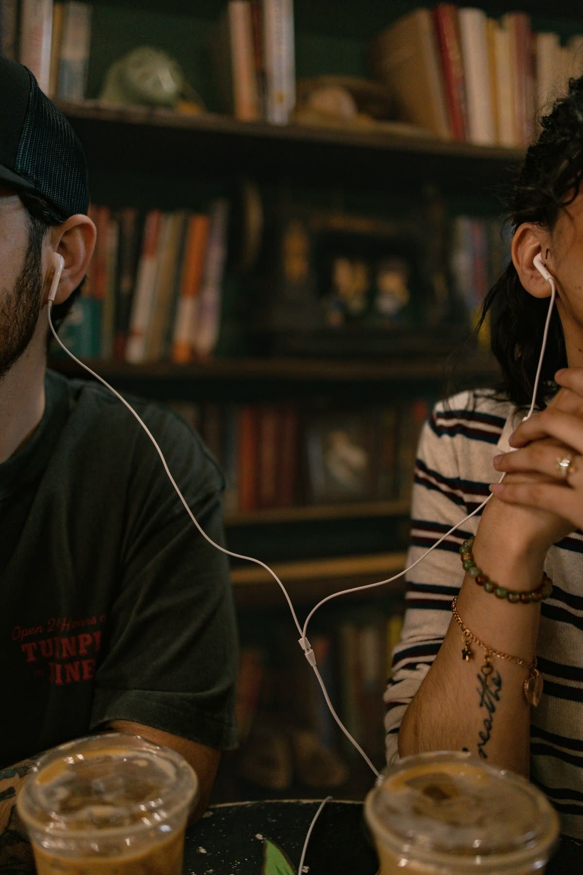 Two people sharing earbuds listening to music together at a cafe table with iced drinks.