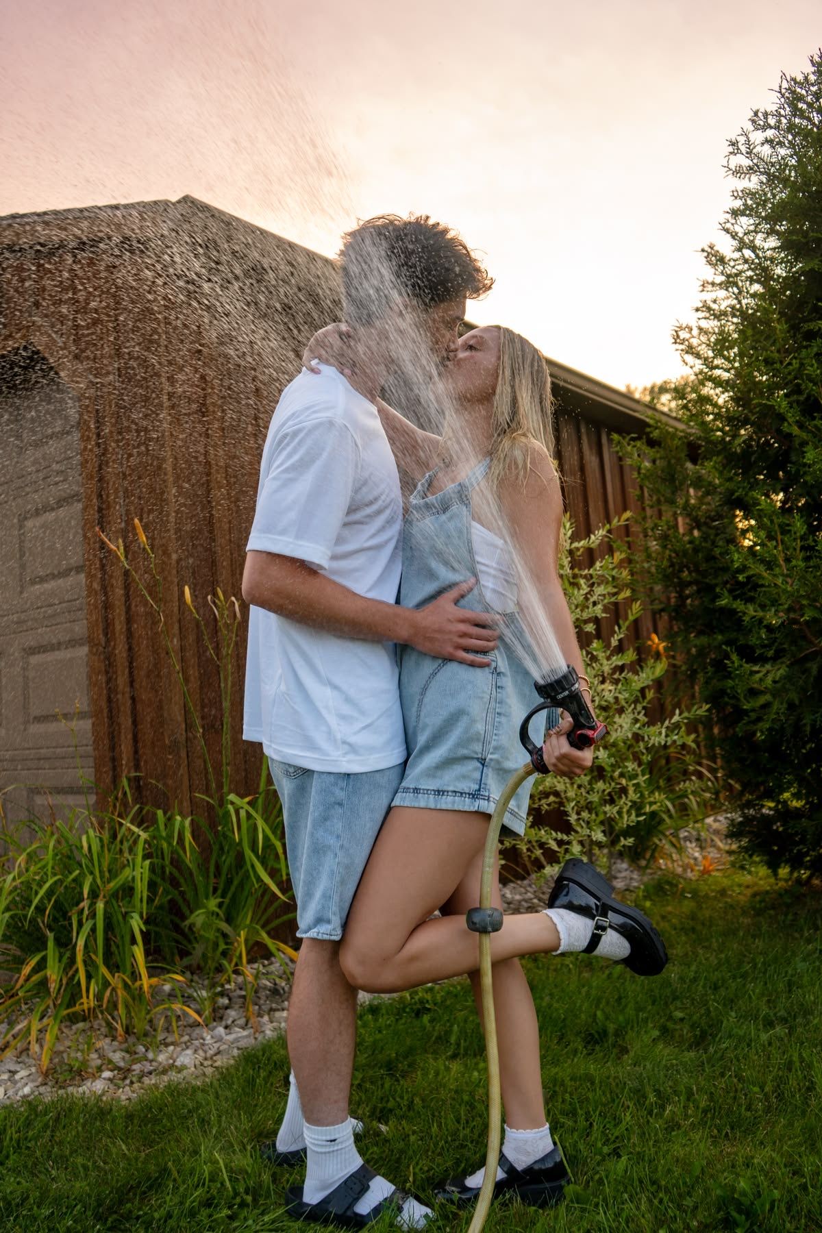 Couple kissing in backyard while being sprayed with garden hose at sunset.