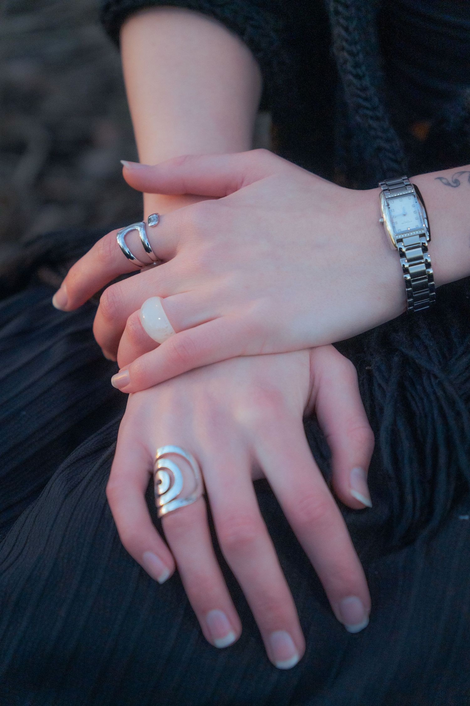 Close-up of two hands adorned with silver rings and a watch resting on black fabric.