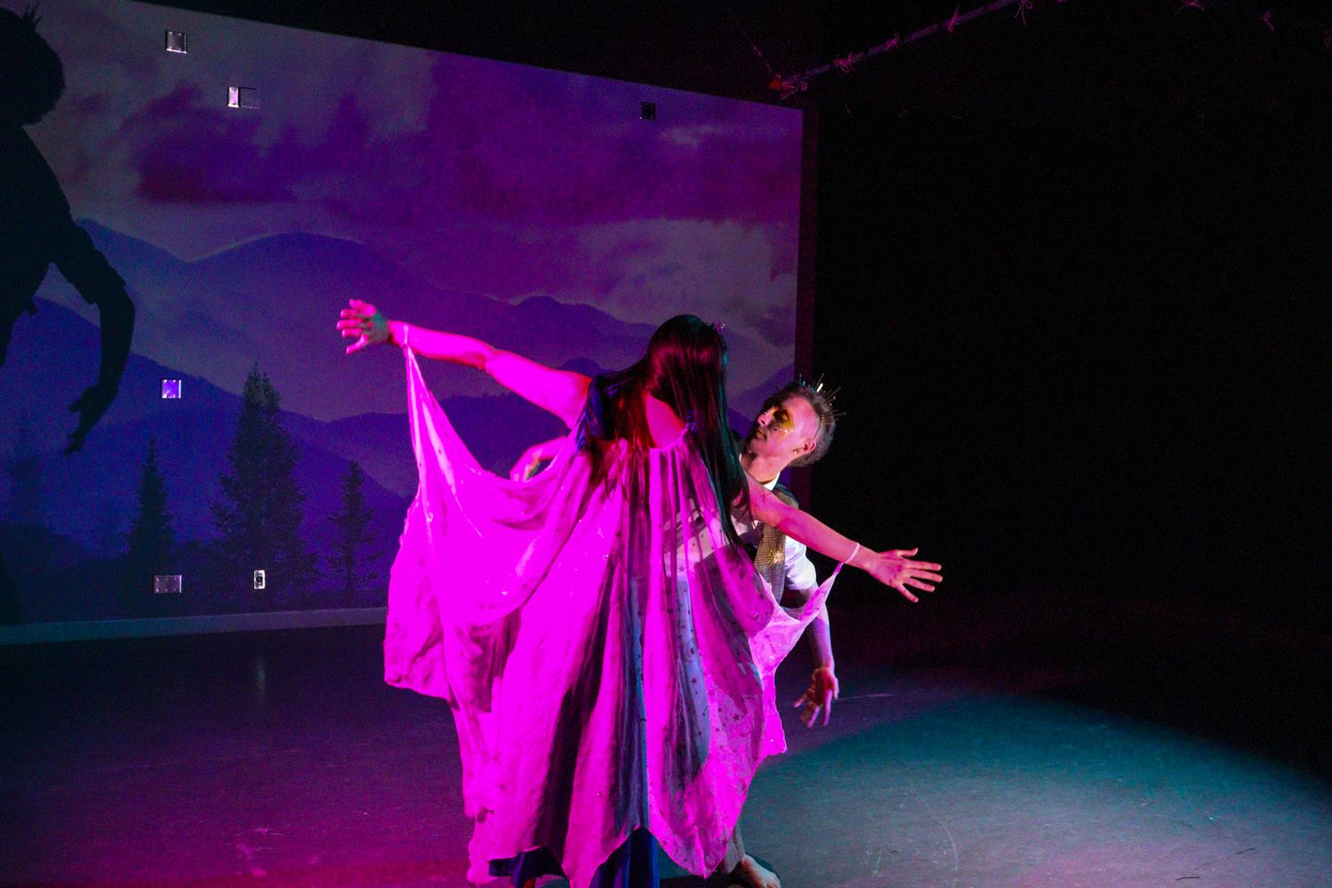 Two performers in flowing pink and iridescent costumes dance on stage with a colorful forest backdrop.