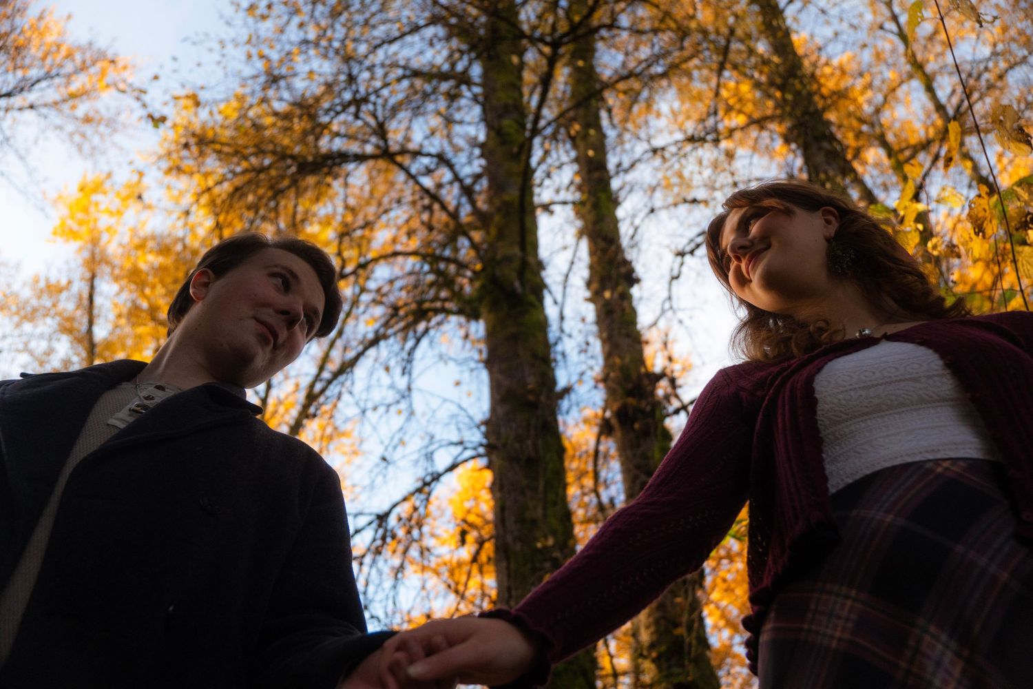 Young couple holding hands looking at each other beneath tall autumn trees with golden leaves.