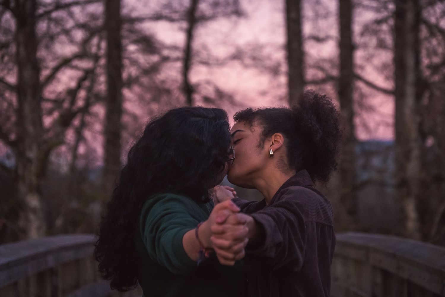 Two women share a romantic kiss on a bridge at twilight, holding hands against a purple dusky sky backdrop.