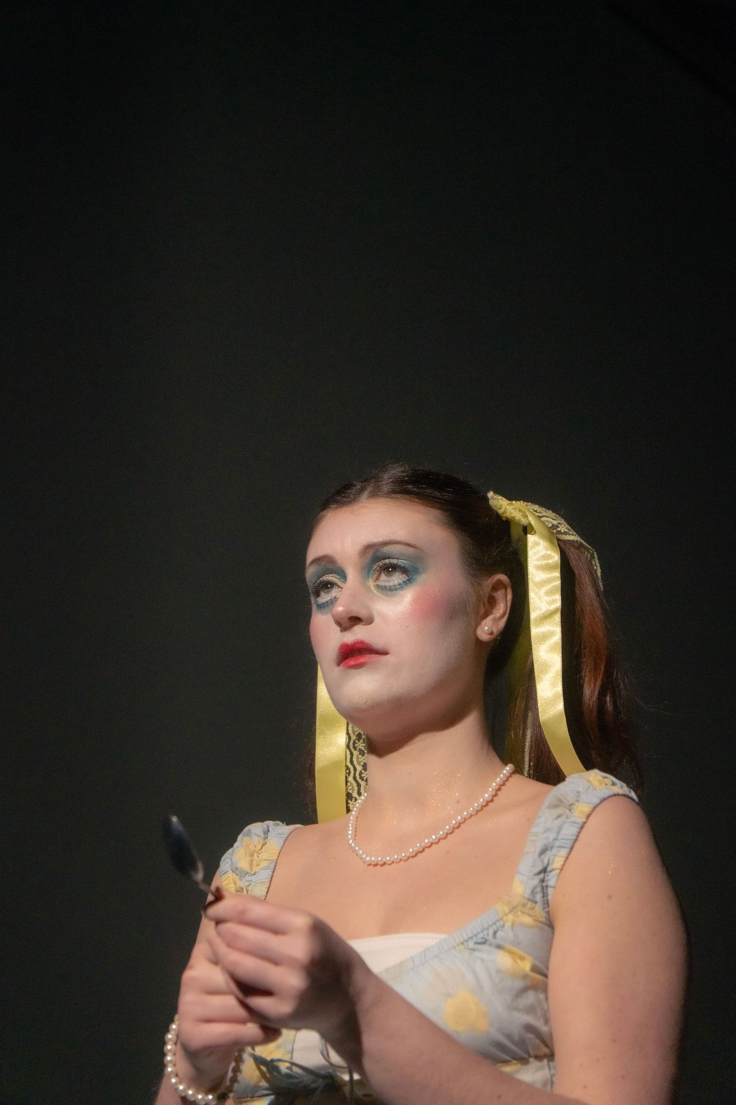 Woman in theatrical makeup with blue eyeshadow, pearl necklace, floral dress, and gold hair bow poses dramatically.