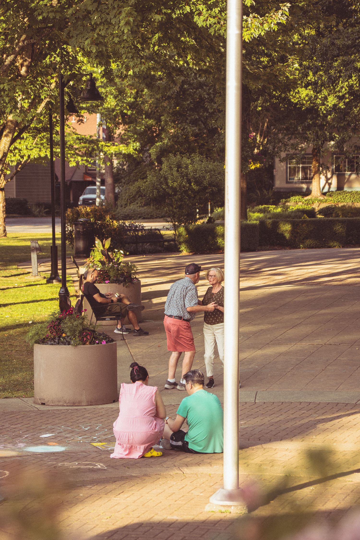 Families enjoying a sunny day in a tree-lined urban park, with children drawing chalk art on brick pavement.