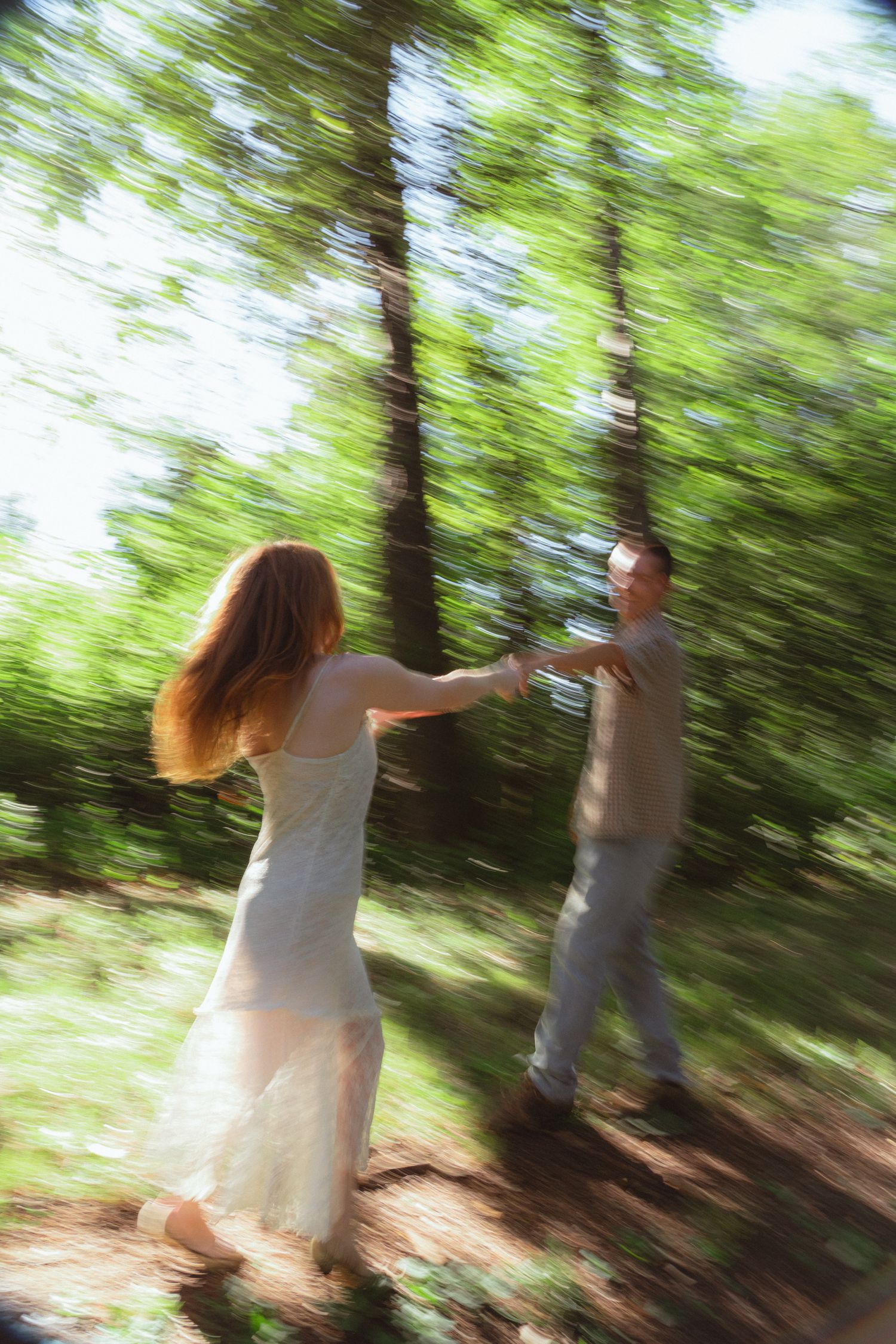A couple dances playfully in a sun-dappled forest, captured with a dreamy motion blur effect.