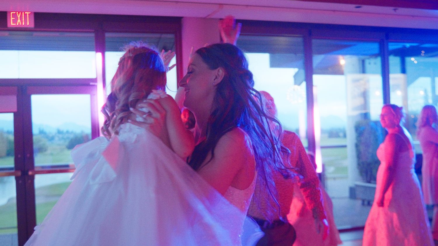 Bride and bridesmaid share an emotional hug on the wedding dance floor under colorful pink and blue lights.