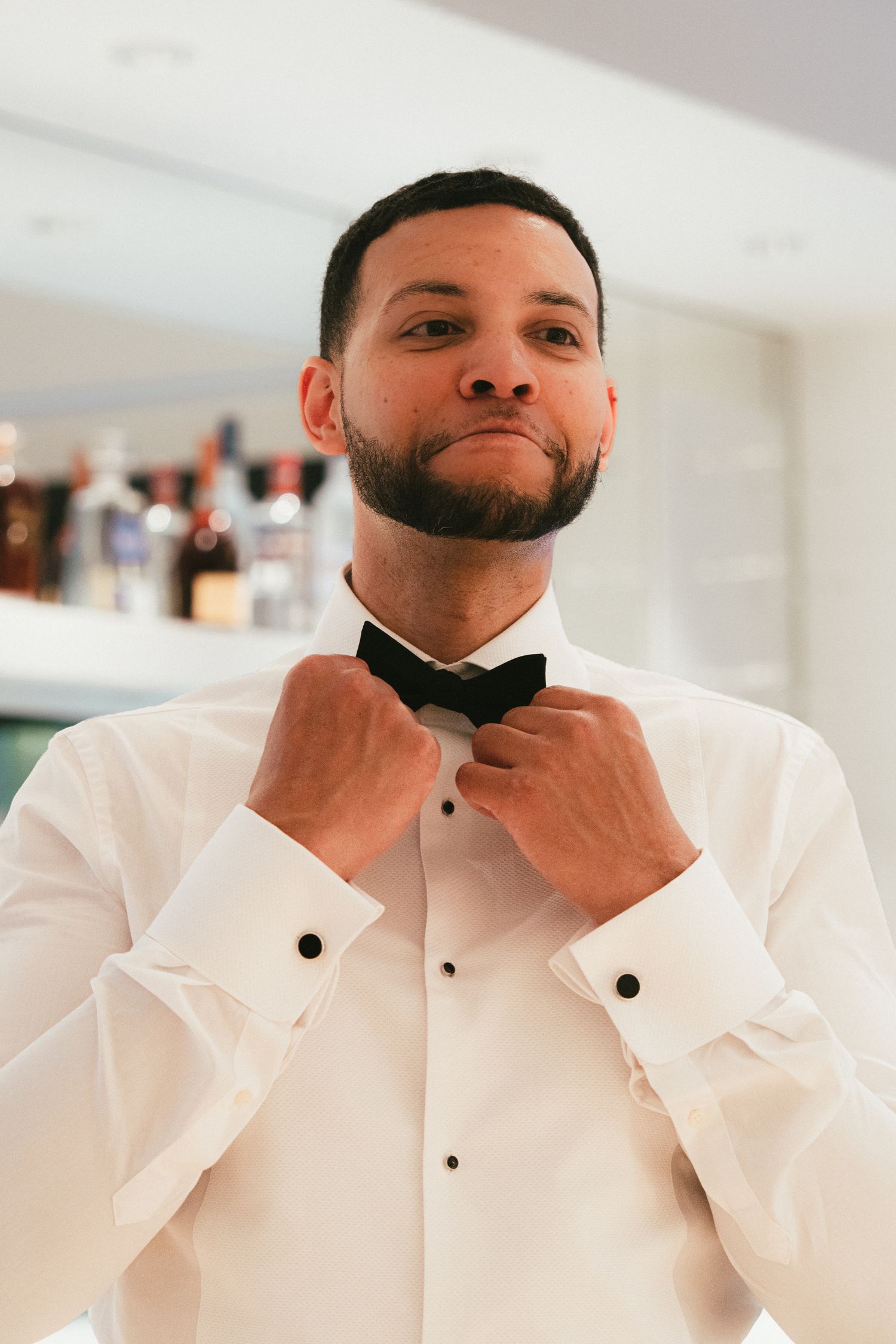 Handsome groom in white tuxedo shirt adjusting his black bow tie while getting ready for the wedding.