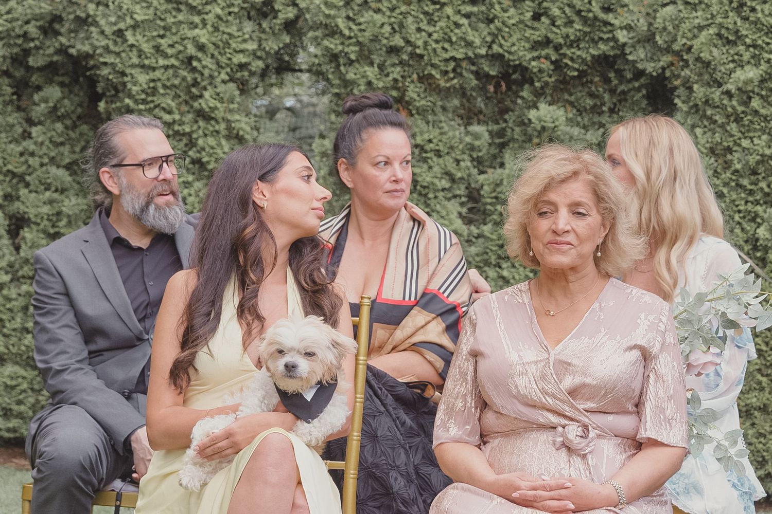 Group of five people sitting outdoors with a small white dog, dressed in elegant attire for a formal occasion.