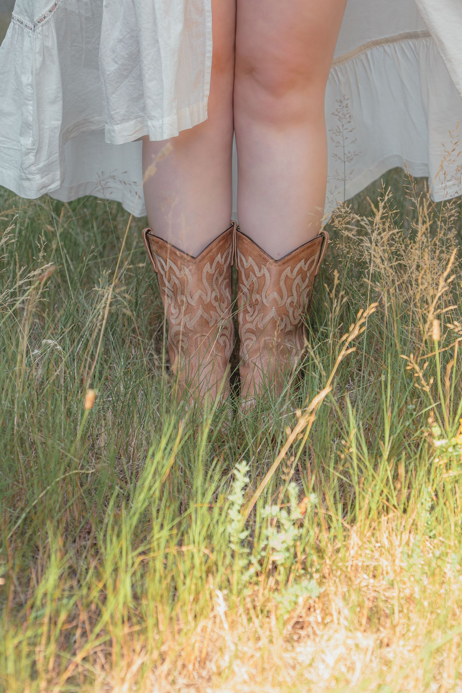 Person wearing tan cowboy boots and white dress standing in tall green grass field.