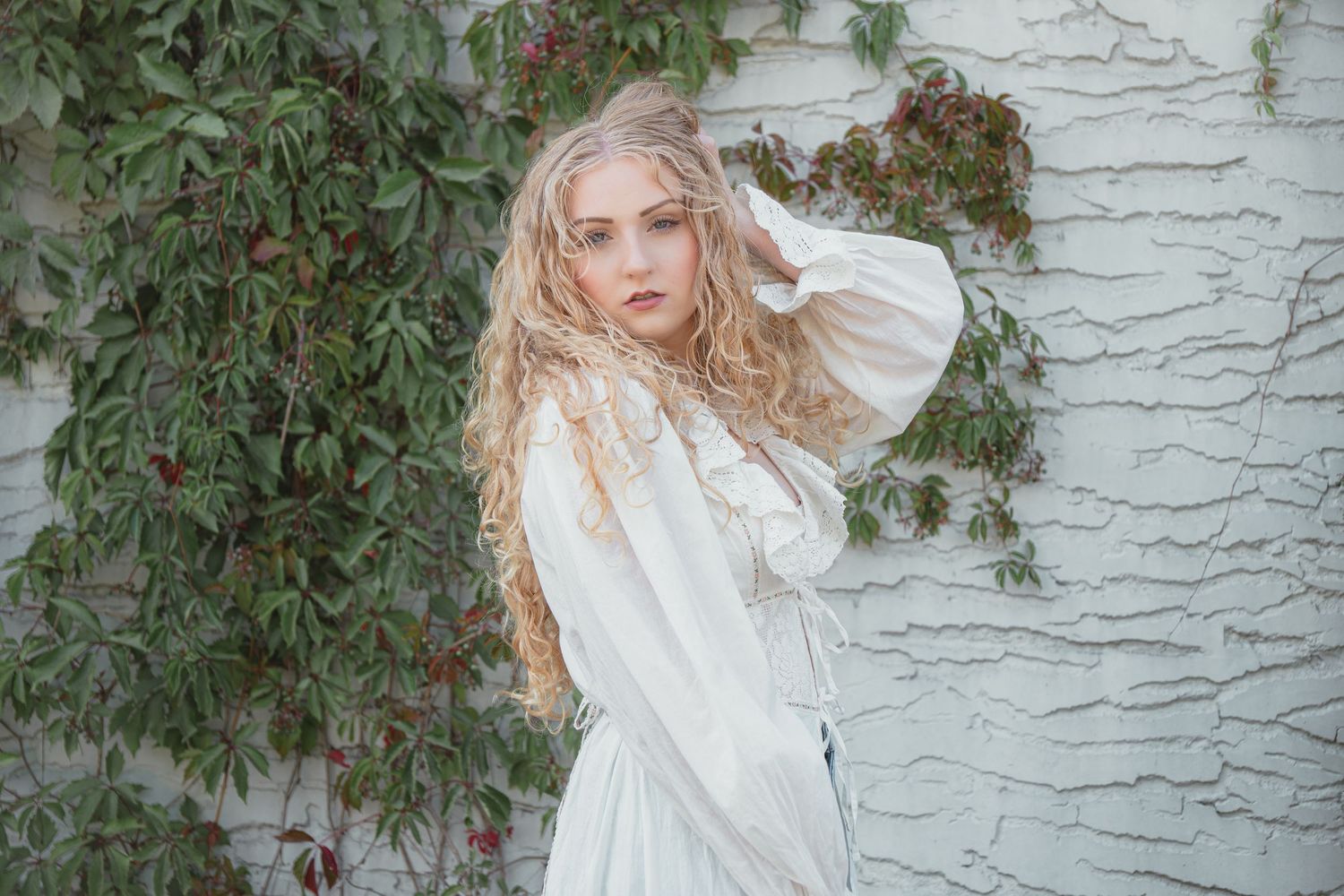 Young blonde woman in white lace dress poses gracefully against a white textured wall with green ivy.