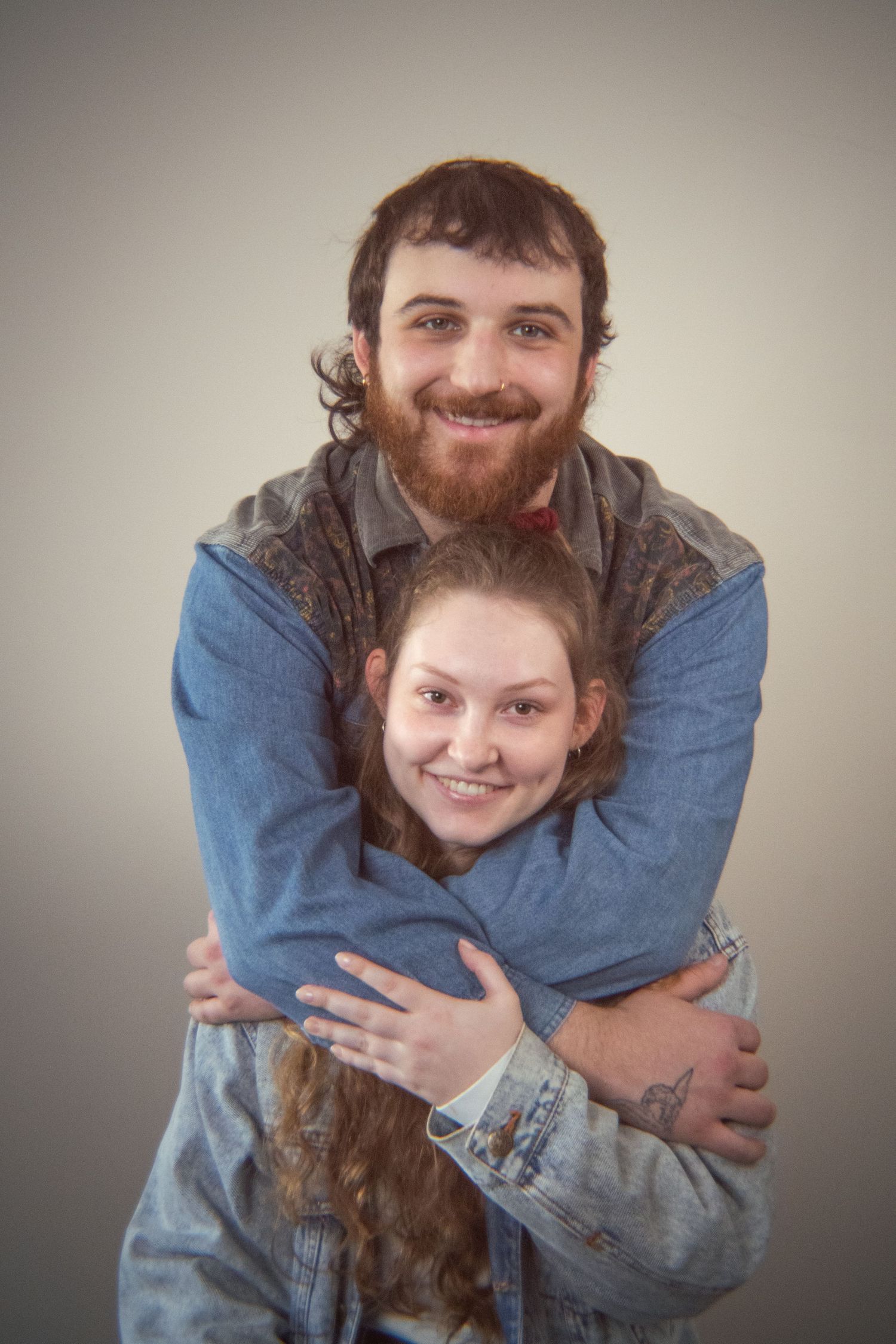 A smiling bearded man wraps his arms around a young woman in a denim jacket in a playful hug.