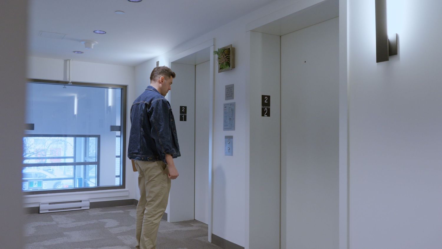 Man in denim jacket standing near elevator doors in a modern building hallway with large windows.