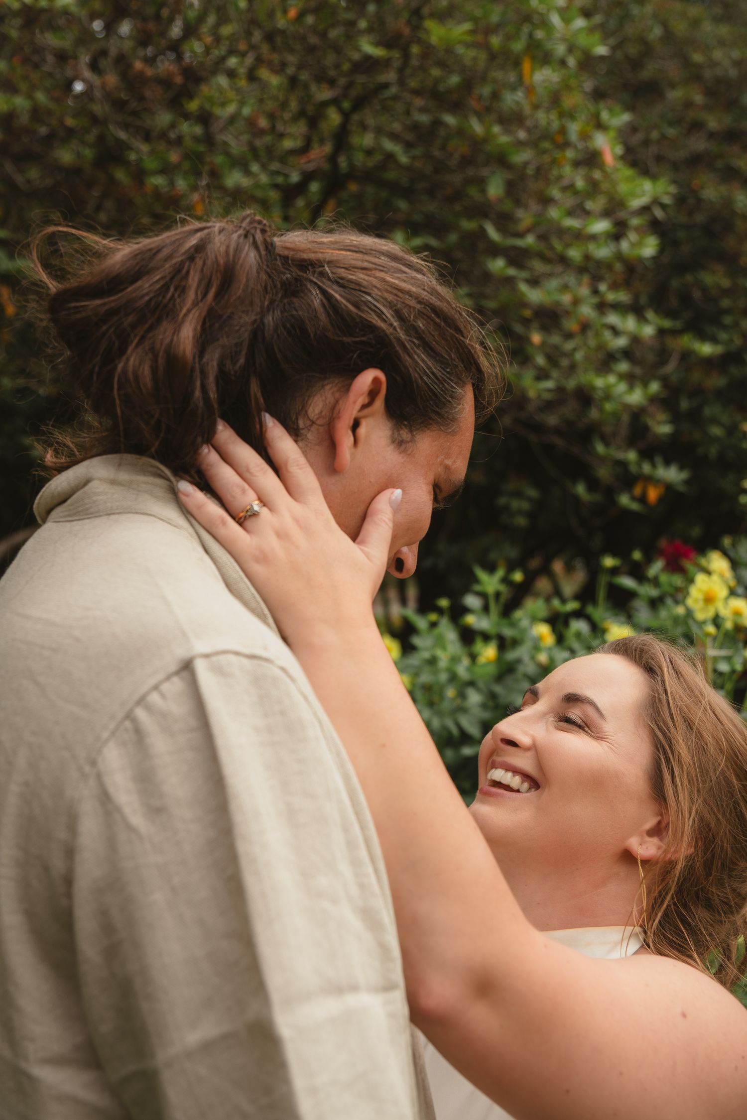 Bride laughs joyfully as she tenderly holds groom's face close in an intimate outdoor garden moment.