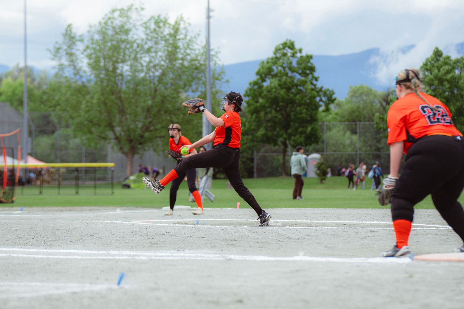 Women's softball pitcher delivers a pitch during an outdoor game with mountains visible in the background.