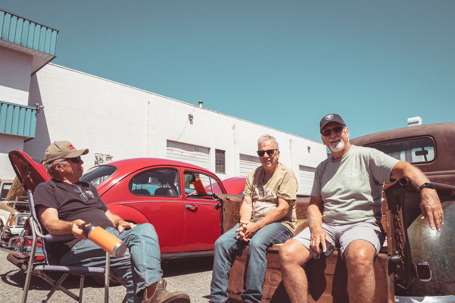 Three men relaxing outdoors at a classic car show with a vintage red Volkswagen Beetle in the background.