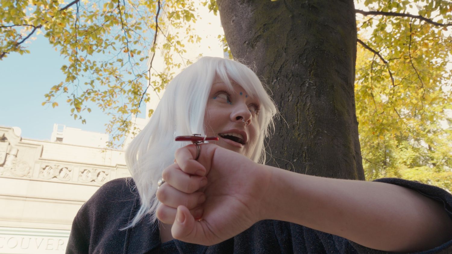 Woman with white hair holding a small object outdoors near autumn trees.
