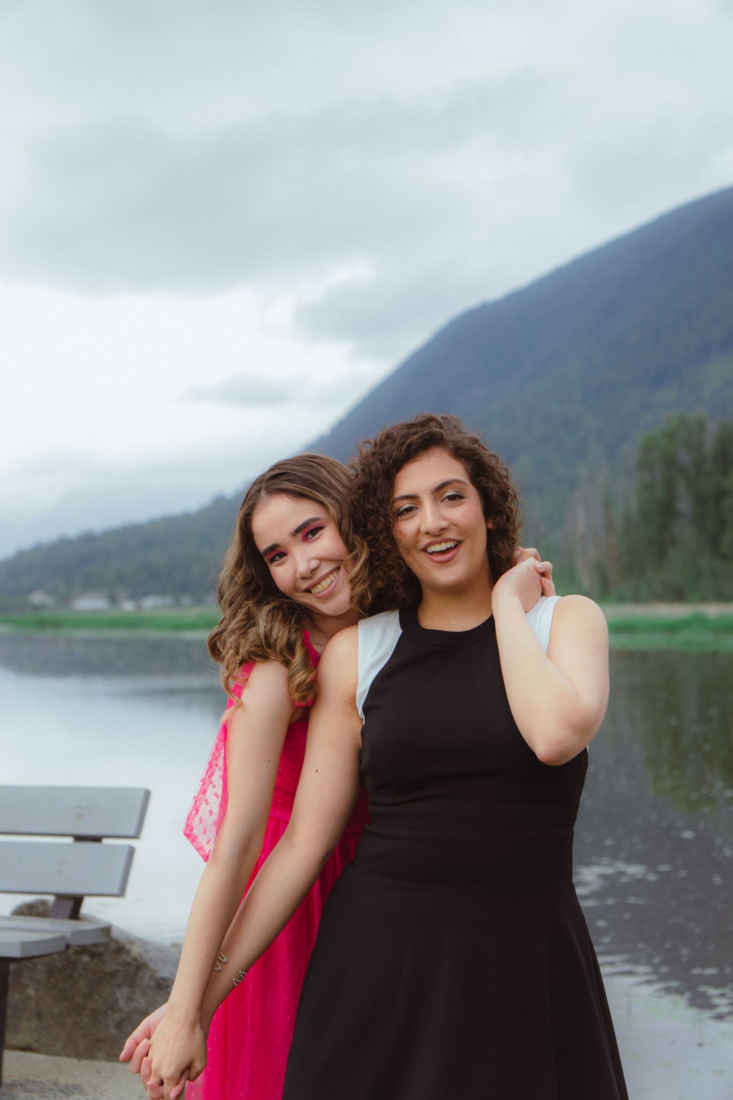 Two smiling women pose together near a mountain lake, one in pink and one in black dress.